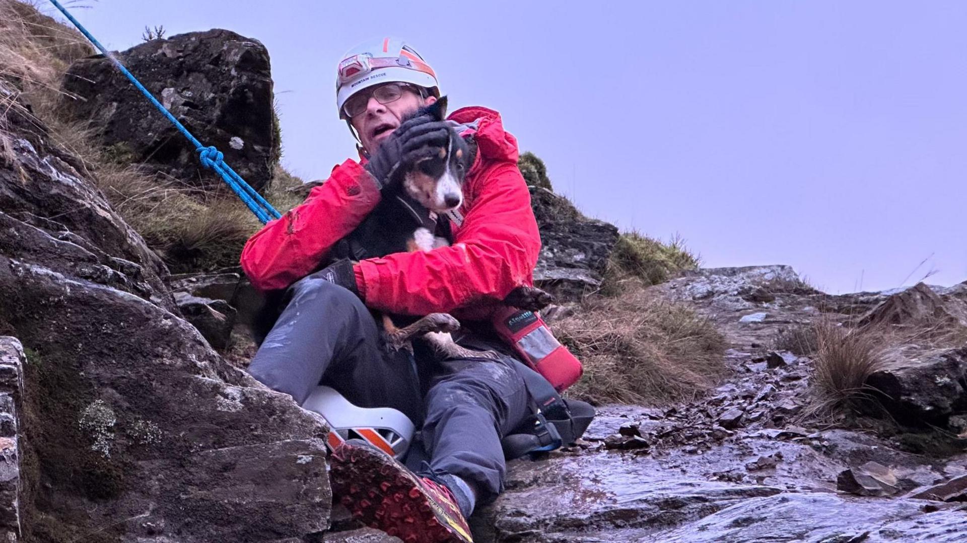 Man in helmet and red jacket holding a dog in his arms. They are on the side of a mountain in the mud. 