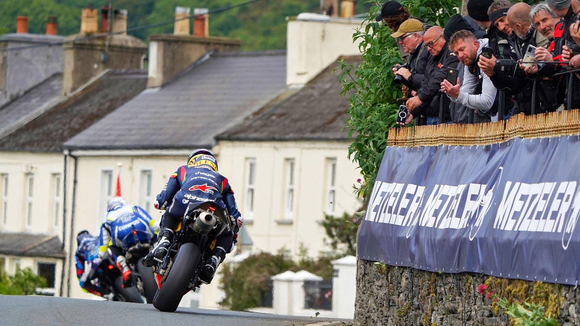 Three TT bikes riding away, with a row of houses in front of them and fans leaning over a stone wall to the right.