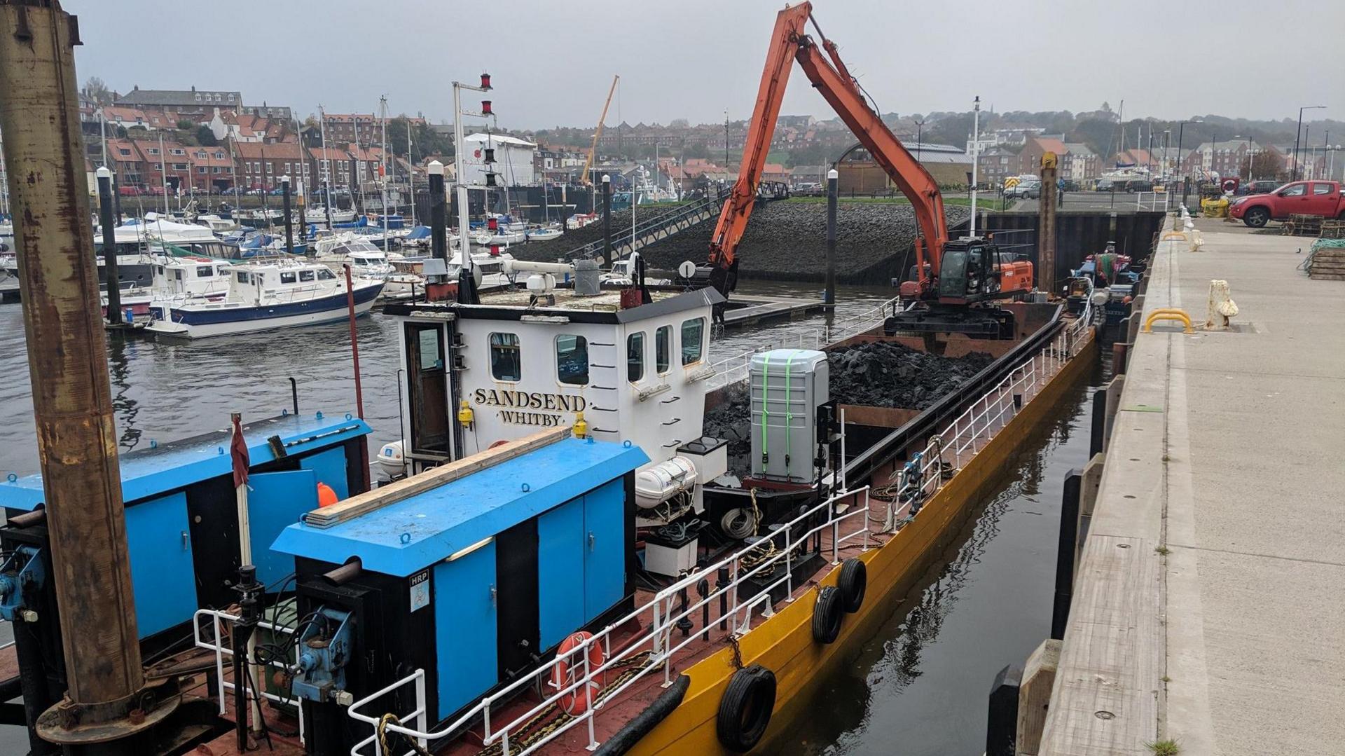 A dredging vessel alongside a harbour painted in a mix of blue, white and yellow, with the name Sandsend written on the white cabin.