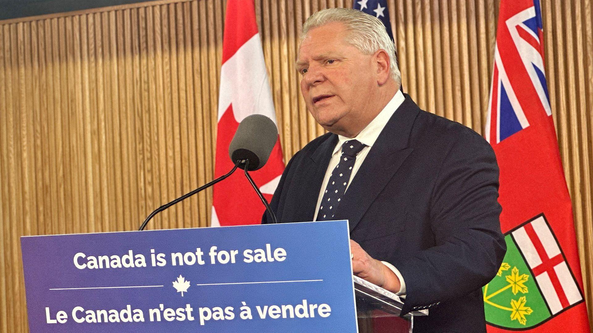 Doug Ford standing front of a sign saying 'Canada is not for sale' while he speaks into a microphone