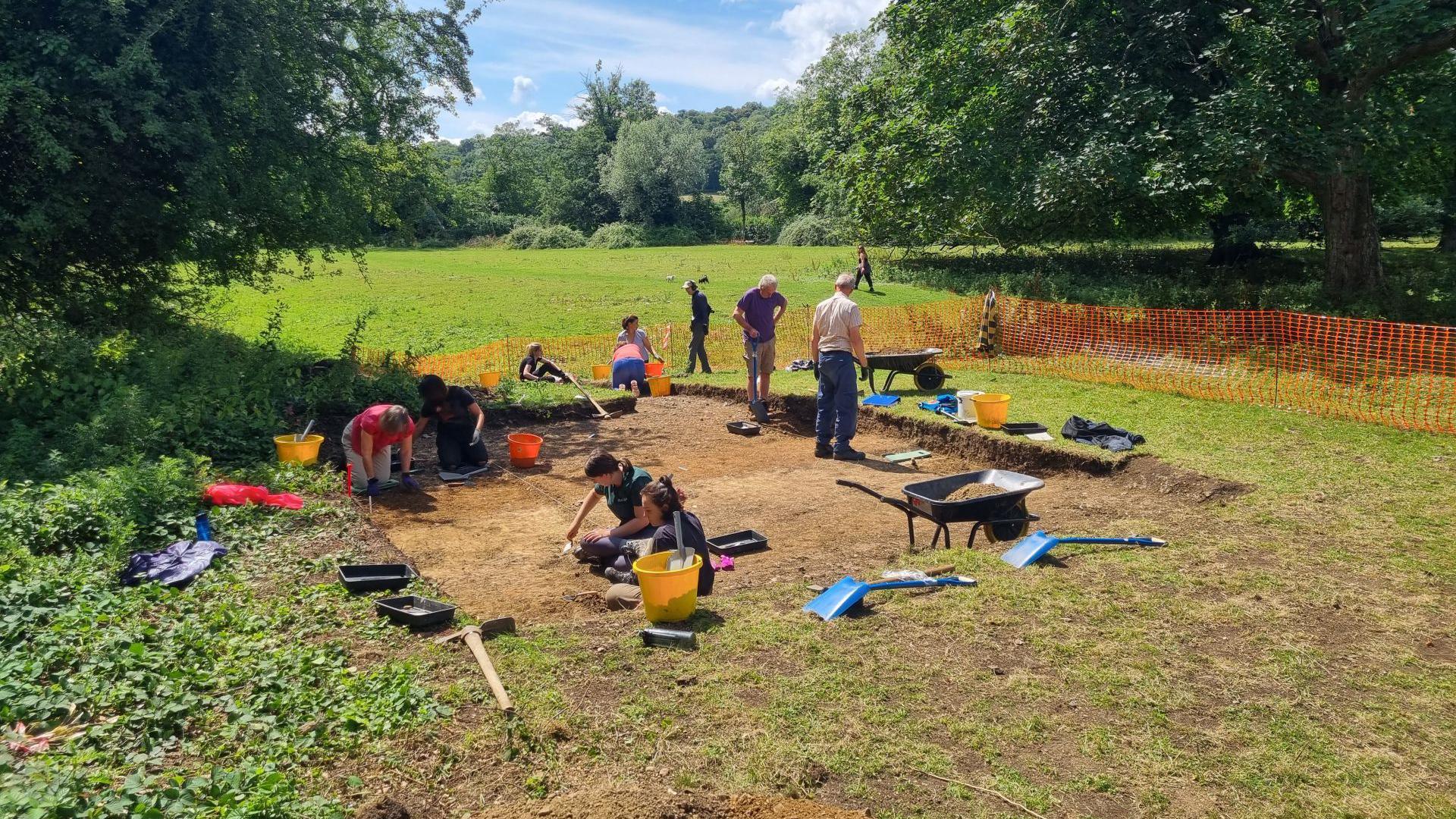 A team of archaeologists works in a hole in a grassy field. It is sunny. 