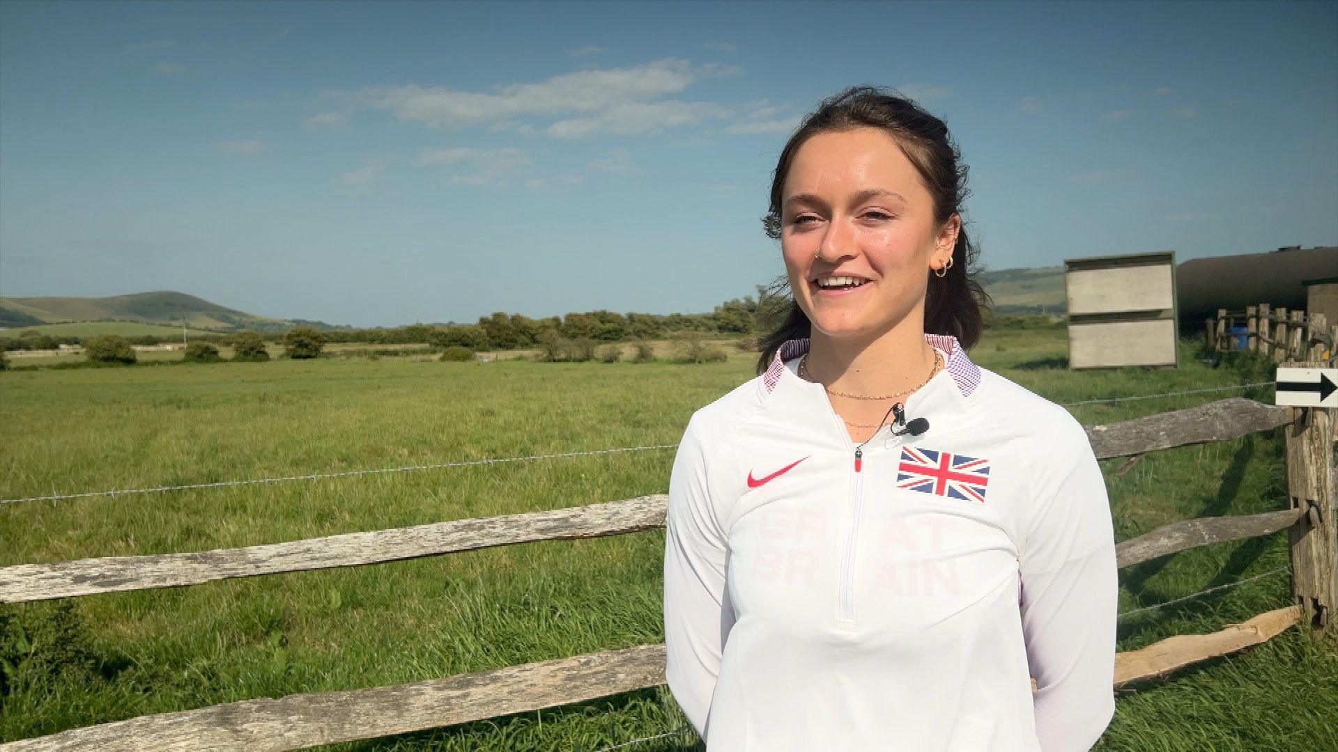 Gemma Tutton, wearing a white GB athletics top, stands beside a farm gate, with a field in the background.