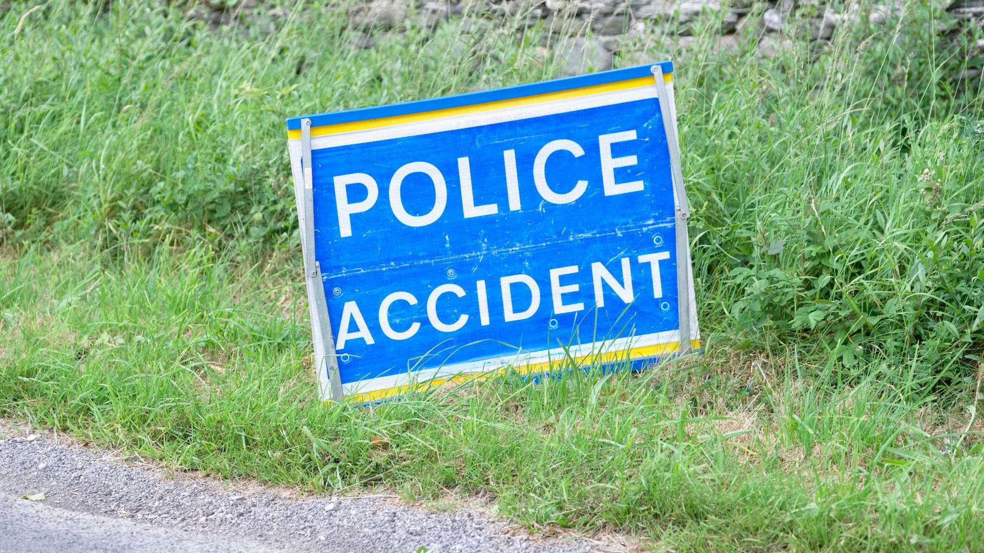 A police accident sign with white lettering on a blue background on a grass verge near a road