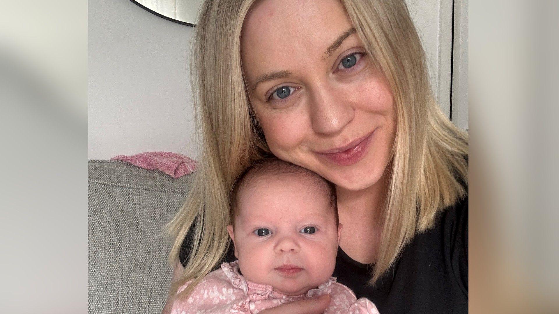 A woman with mid-length straight blonde hair wearing a black top sitting on a sofa holding her baby who has a small amount of dark hair and is wearing a pink onesie.