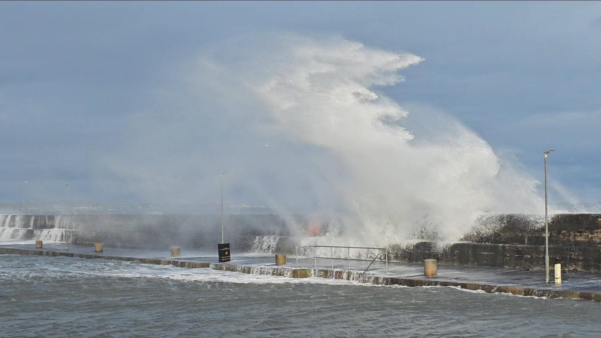 A high wave rushing over the edge of a pier and water is pooling over the wall. The street is flooded with sea water.