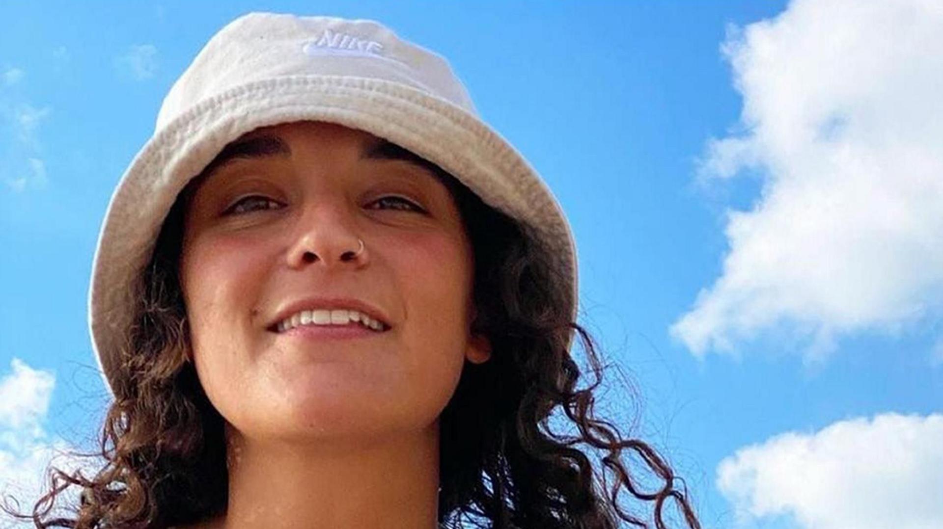 Emily Damari, a young woman with curly dark brown hair wearing a white bucket hat, smiles down at the camera from above, with a blue sky behind her.