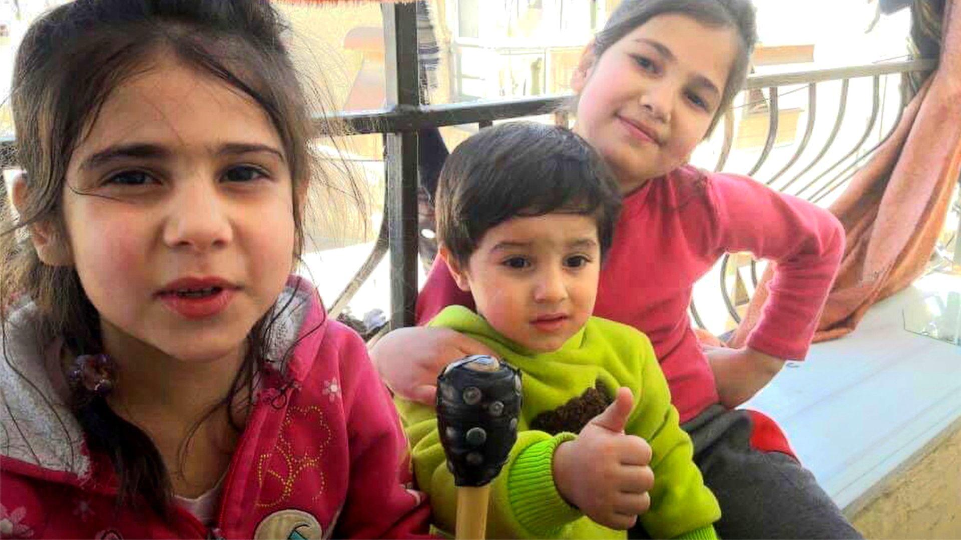 Two little girls and a little boy with dark hair and in bright tops smile for the camera 
