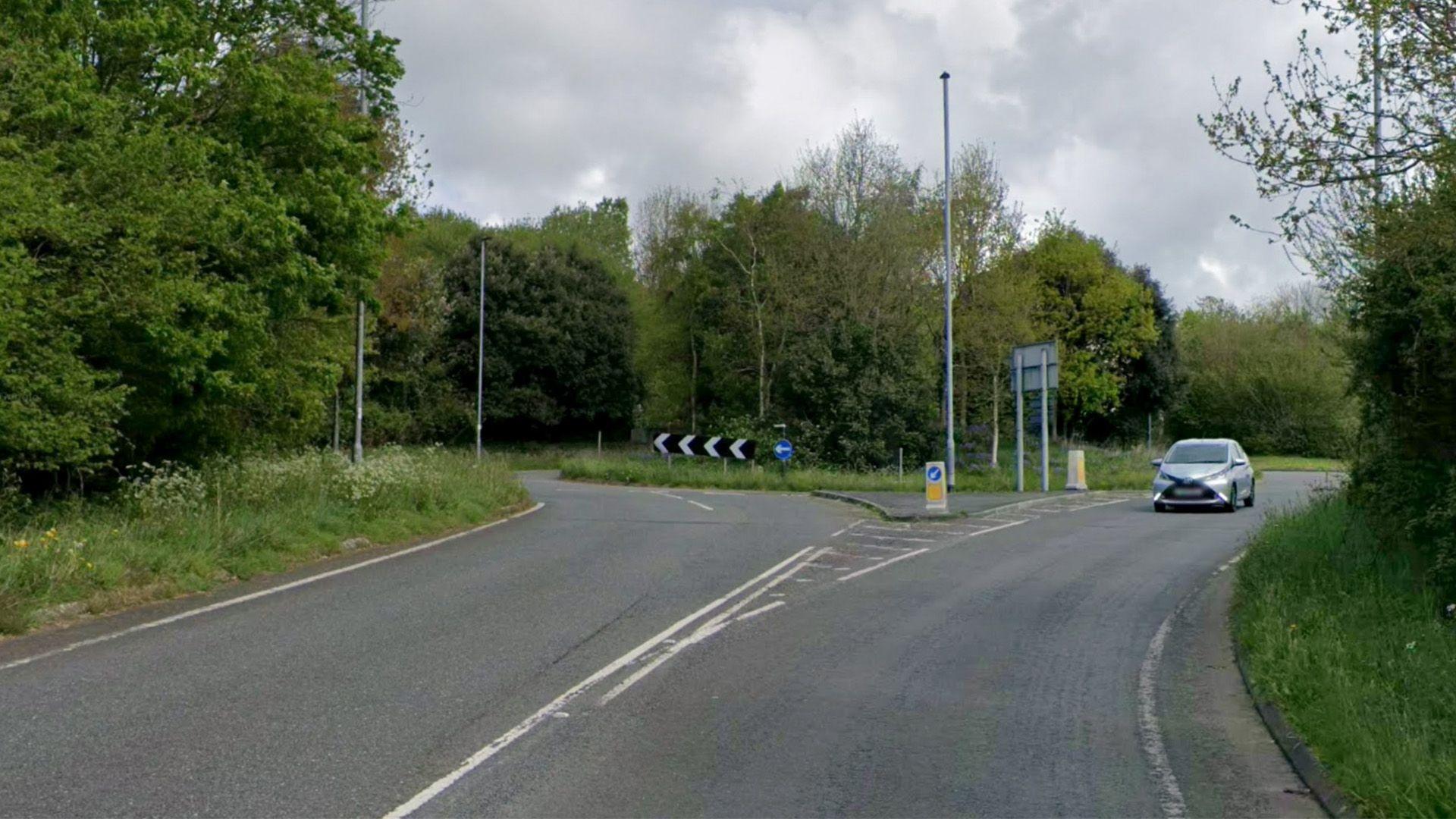A google maps grab of a road, leading up to a roundabout. One silver car is visible. There is greenery on either side and in the centre of the roundabout.