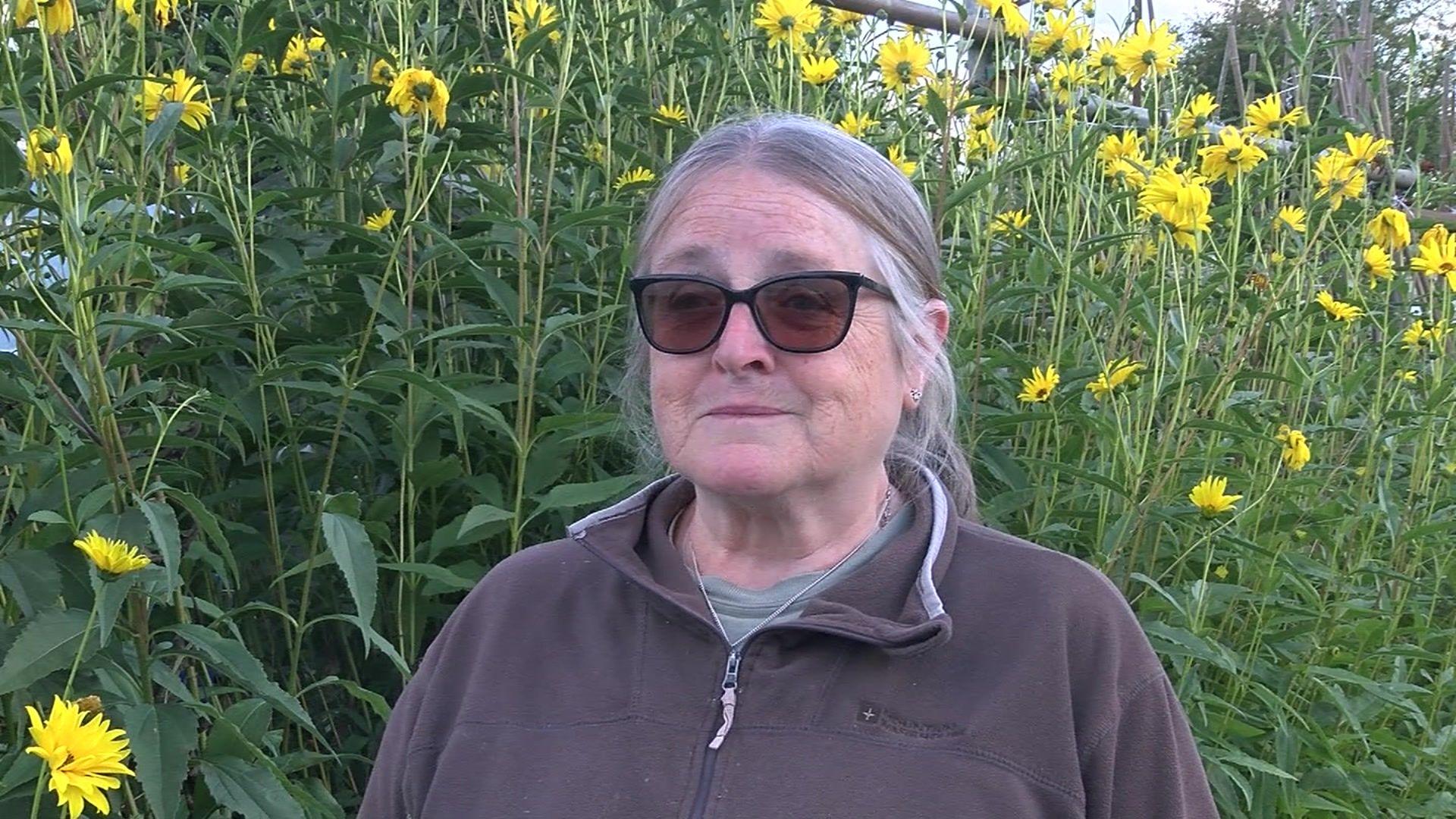 Jenny Morgan - wearing a brown fleece and sunglasses. Behind her are wild flowers on a grassy bank.