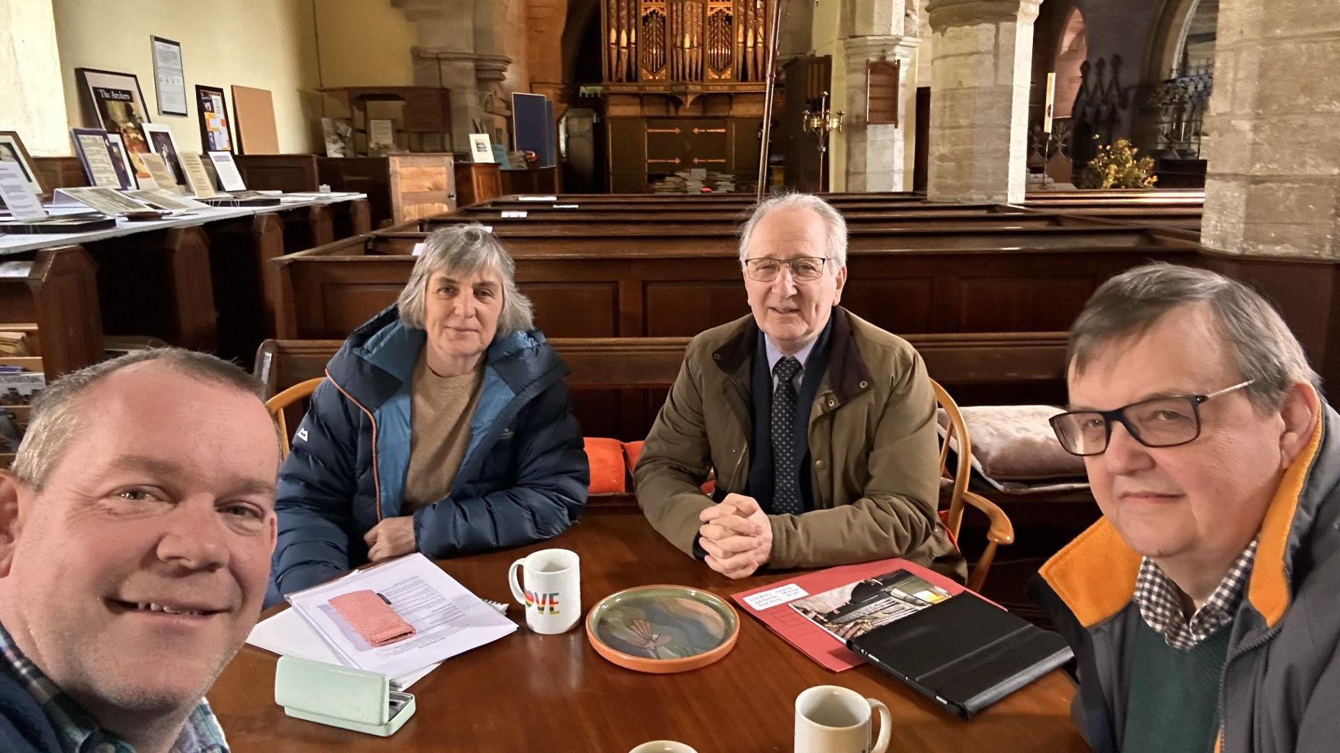 Four people sit at a table in a church, with pews and an organ in the background. They have sheets of paper, notebooks and mugs on a table. Three are men, and one a woman. The man taking the picture is smiling and has short fair hair. The collar of his checked shirt can just be seen. The woman has grey bobbed hair with a fringe and is wearing a blue puffer coat with beige jumper. To her right is a man white white hair and glasses wearing a beige jacket and tie. To his right is a man with dark hair and black-framed glasses in a coat with orange collar, green v-neck jumper and blue checked shirt.