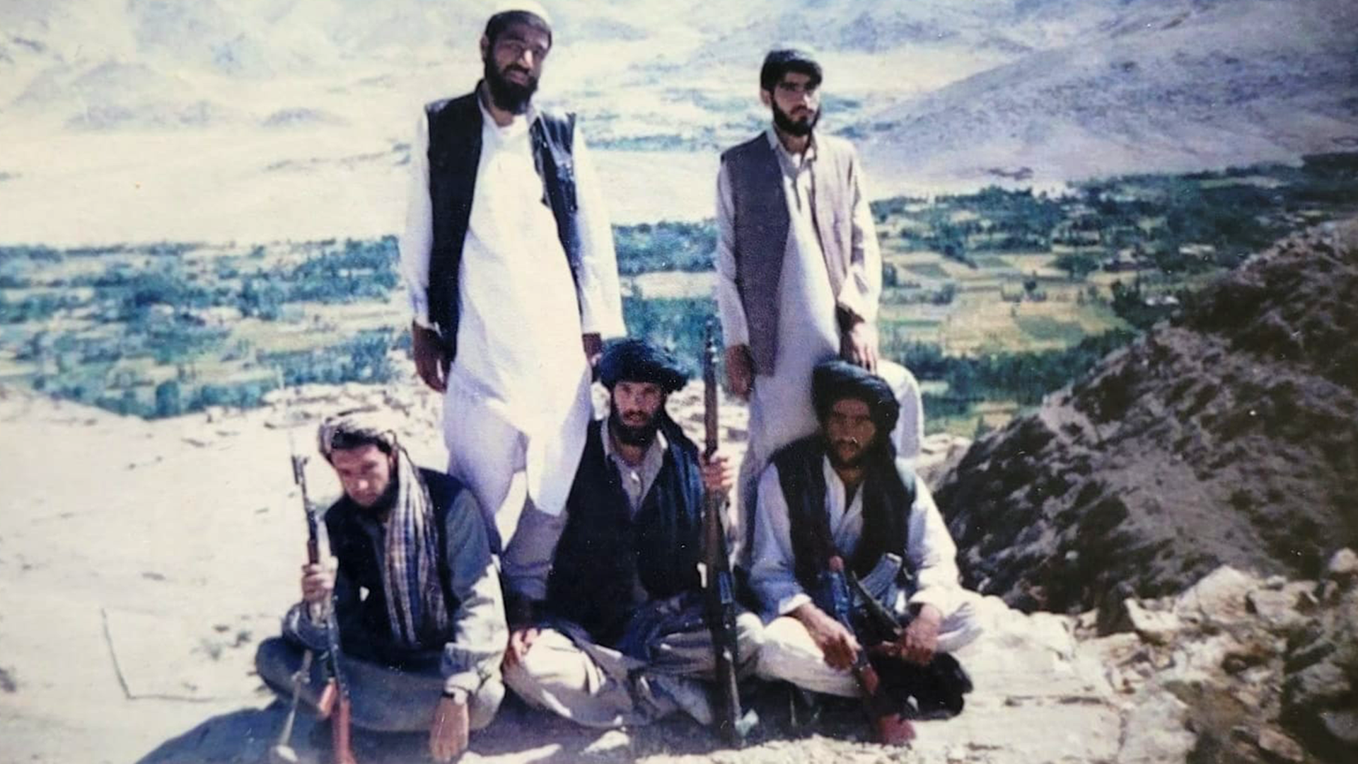 Three men are seated wearing turbans and holding rifles. Two men stand behind them wearing traditional light-coloured tunics, trousers and hats, with dark sleeveless jackets. There are fields and mountains in the background.
