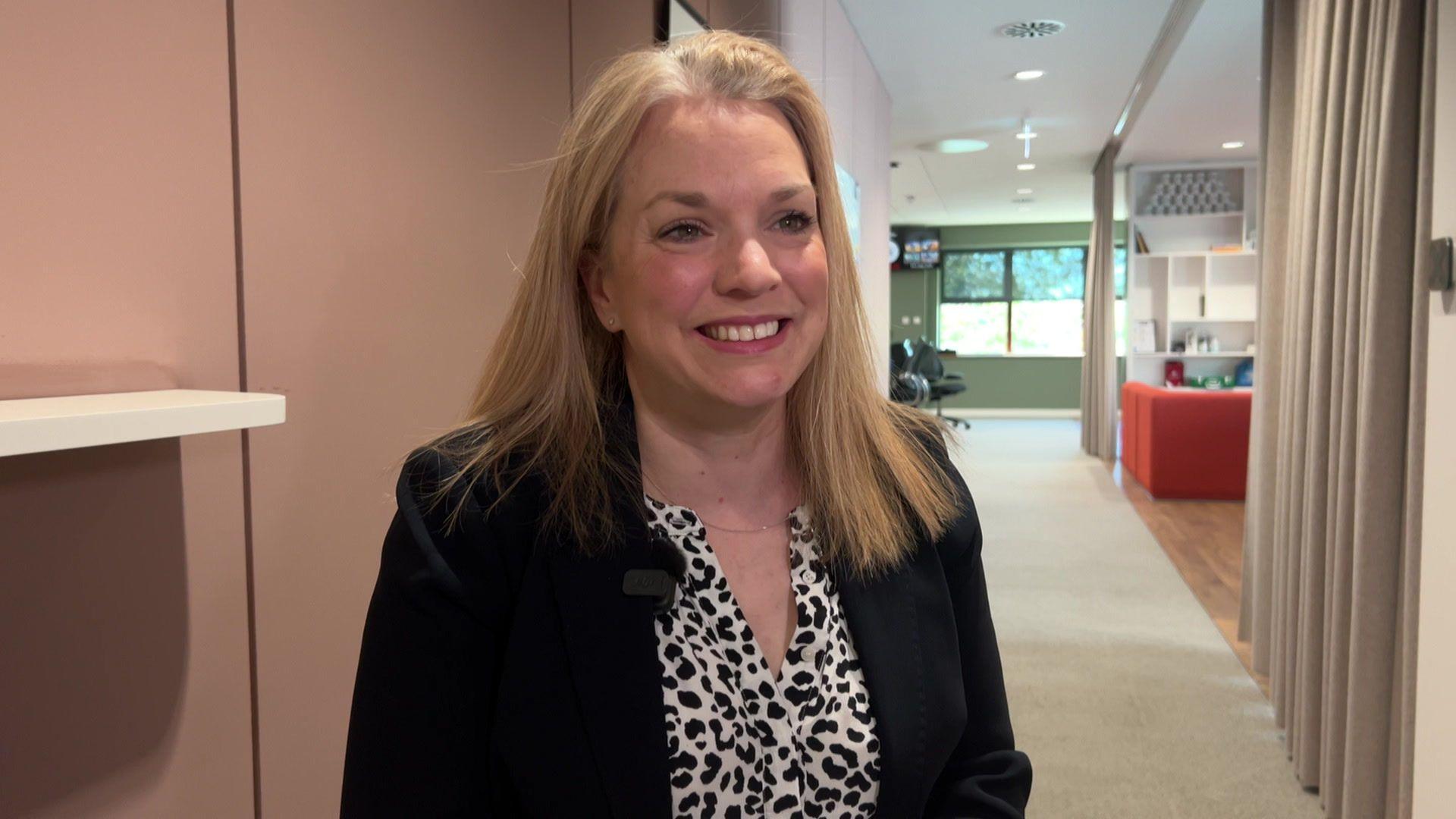 Amy Wallhead smiles at the camera. She is standing in an office, with a pink wall behind her. She has blonde hair and is wearing a black and white blouse and black jacket.