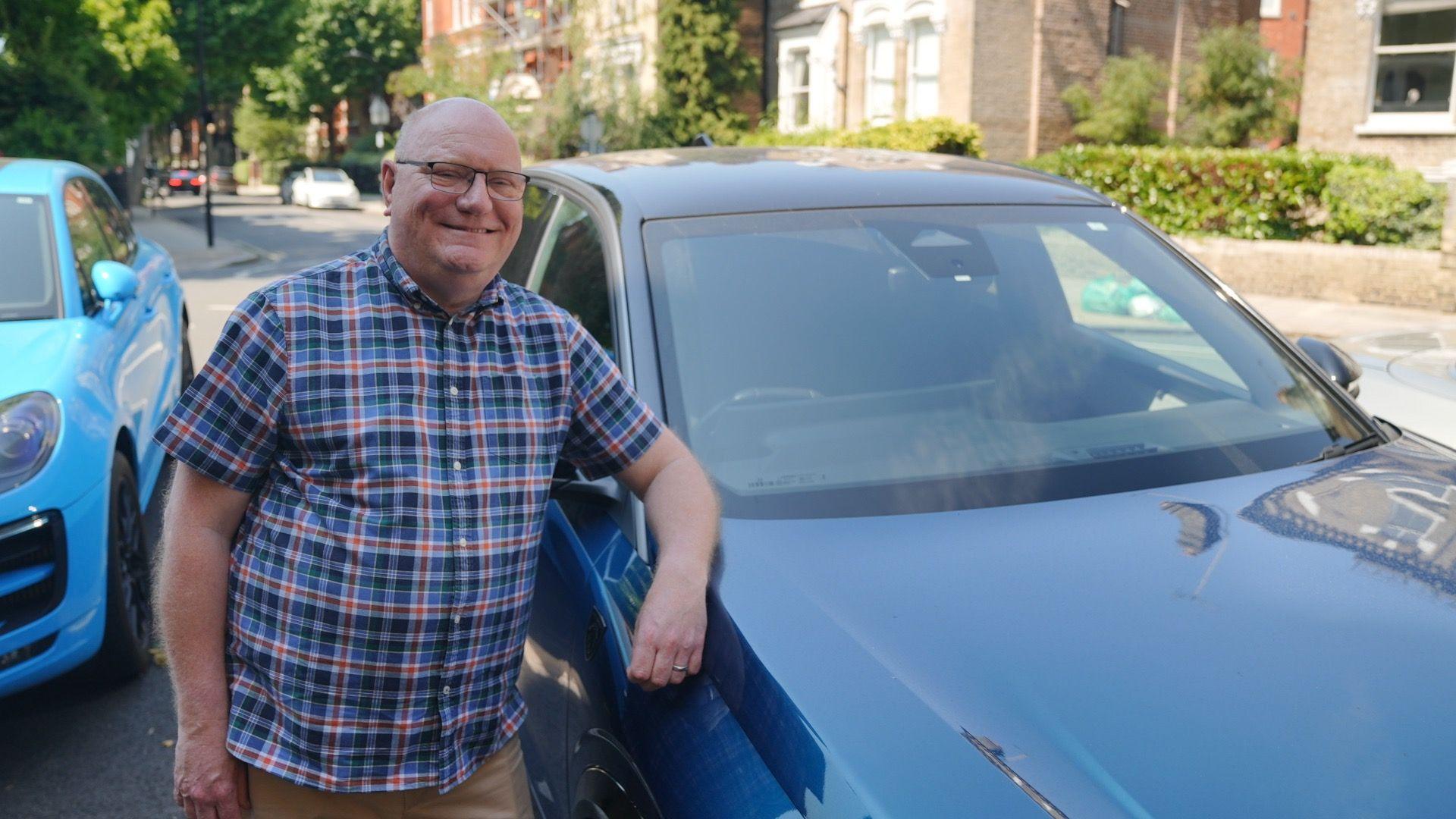 A smiling man in a checked blue and red shirt, and brown trousers leans on his dark blue car parked in the middle of the street. To his right is a lighter blue car and in the background a residential street with other cars driving both ways and front garden hedgerows along the pavement