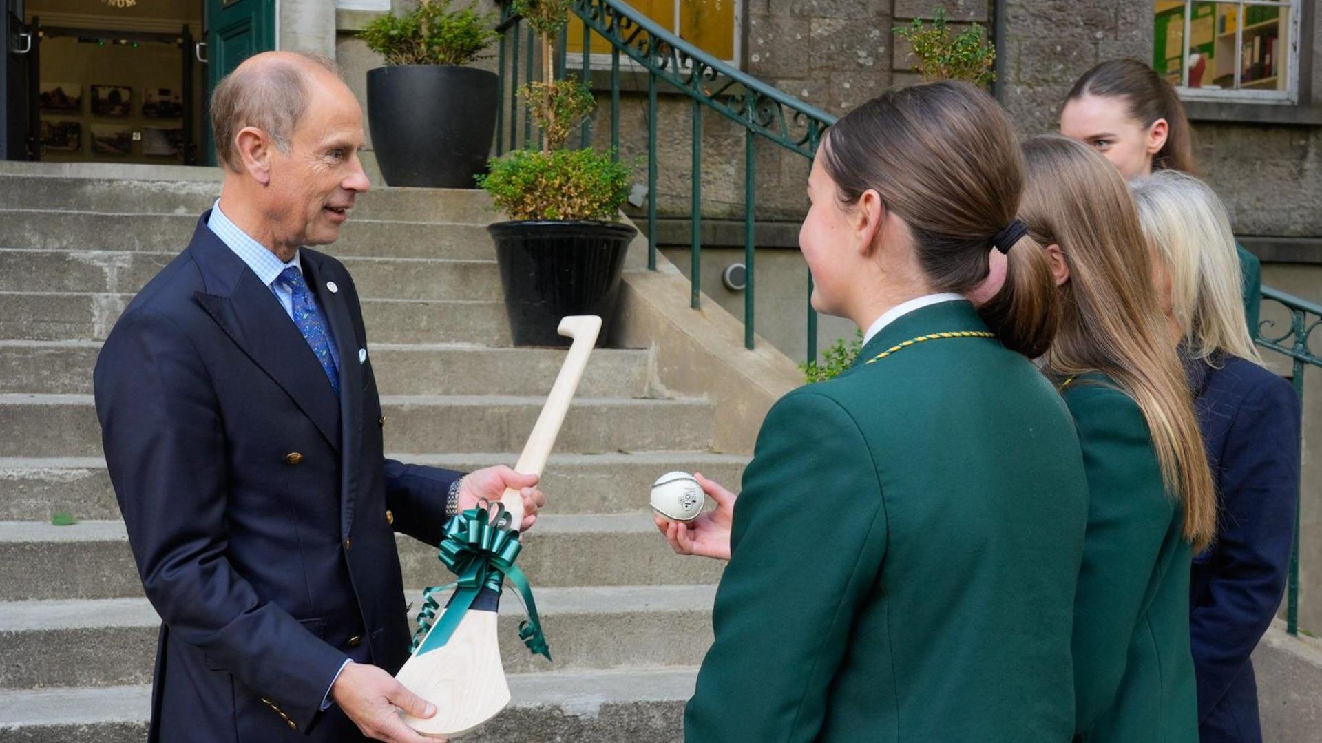 The Duke of Edinburgh, Prince Edward, is being presented with a hurl by a group of pupils from St Catherine's College, Armagh. The hurl has a green bow wrapped around it. 