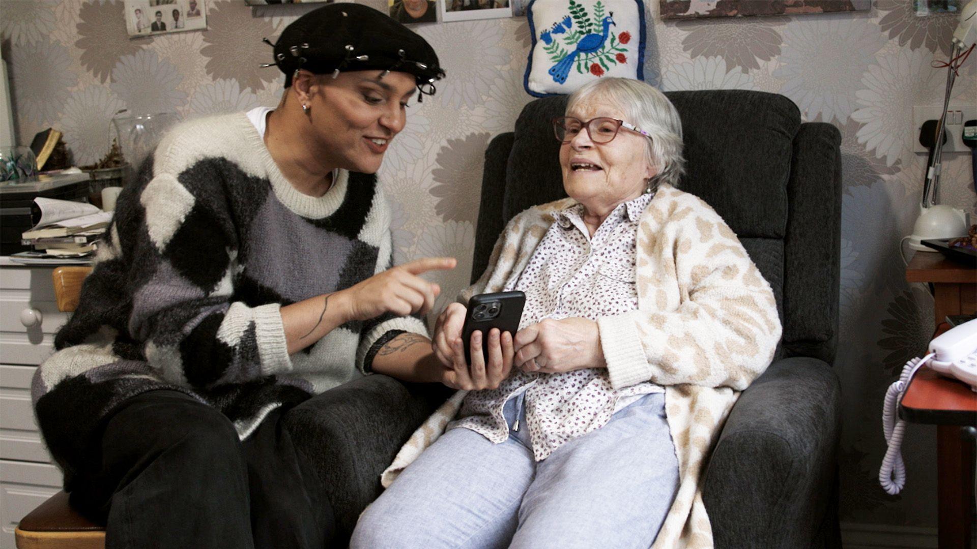 A woman sitting in an armchair is being shown something on a mobile phone by a relative