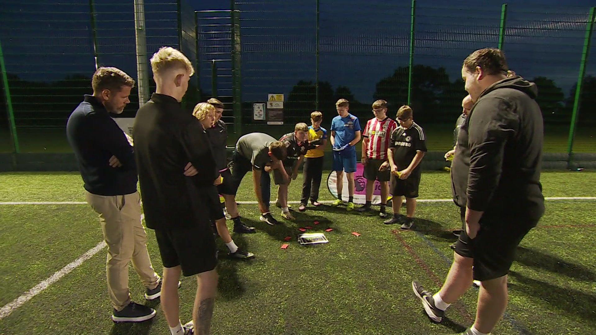 A crowd of young people gathered around, wearing football shirts and dropping red and yellow cards around a photograph on the floor of a green AstroTurf pitch. Footballer James Beattie is on the left and members of both charities are also gathered around.