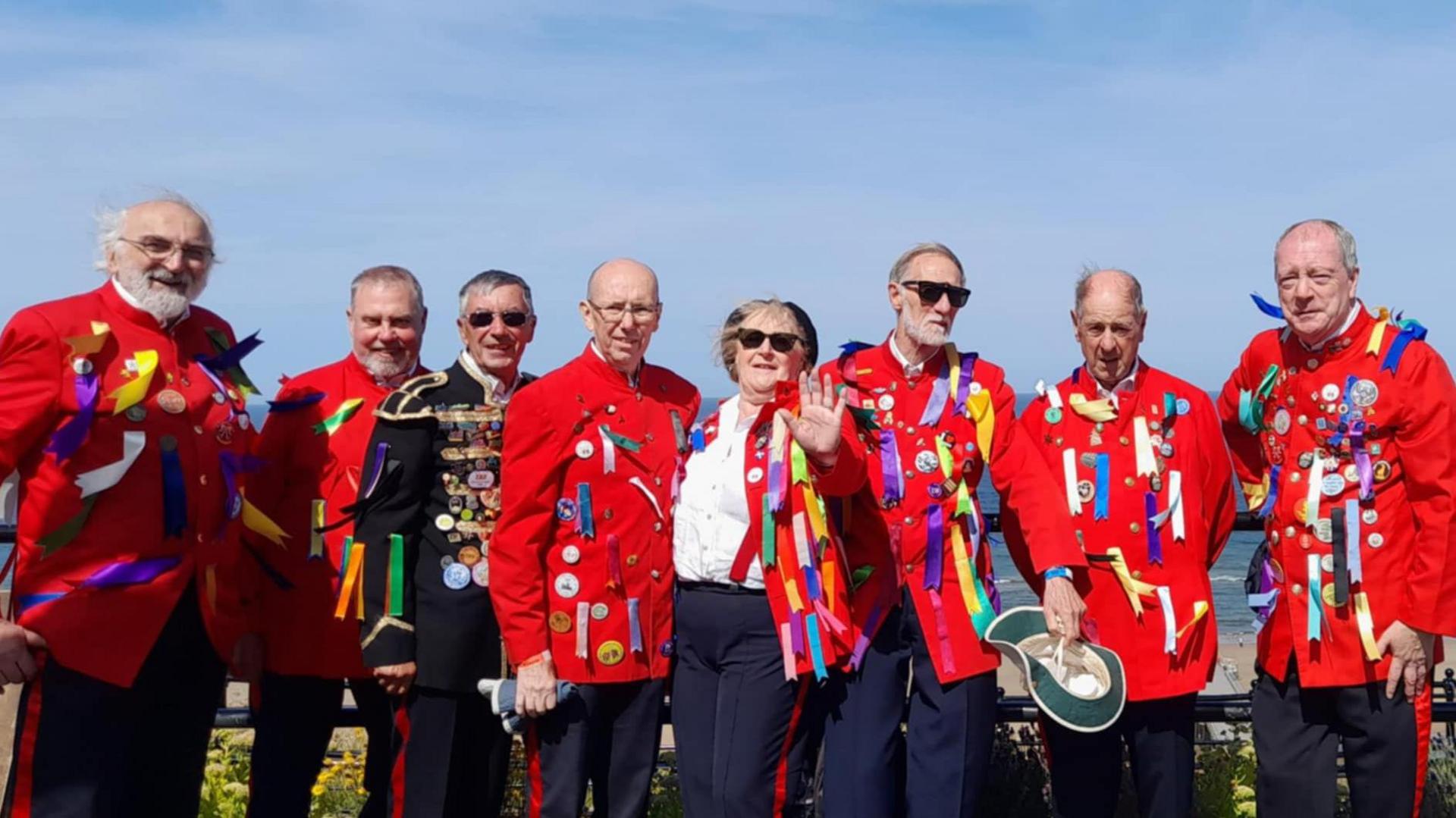 The Redcar Sword Dancers in full red uniform with multicoloured ribbons attached. Eight members of the group are standing in front of a railing at the beach with a blue sky behind them.