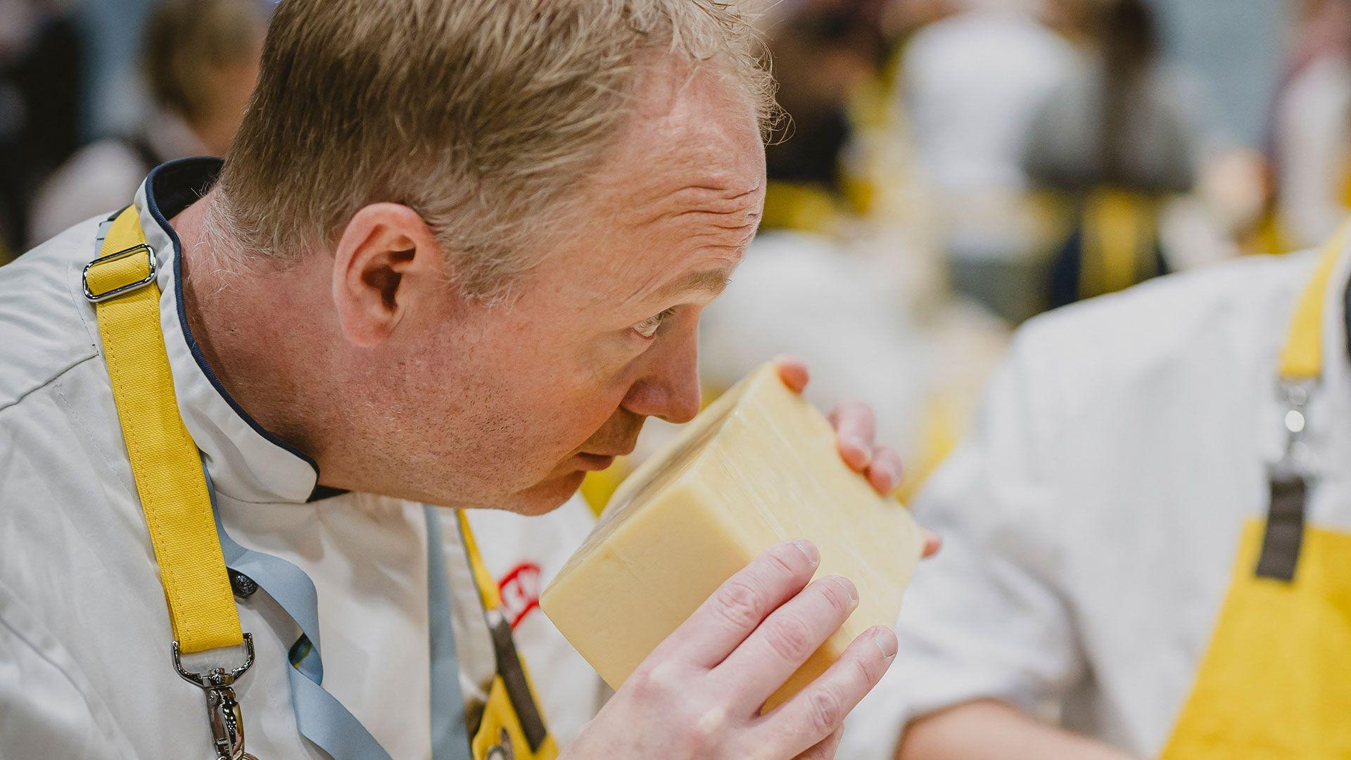 A person in a yellow apron smells a big chunk of cheese