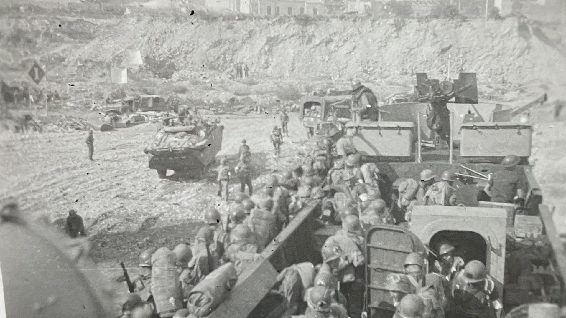 A black and white photo of American troops making their way off a large landing craft wearing full military kit. The photo was taken by the ships commander from the quarter deck.