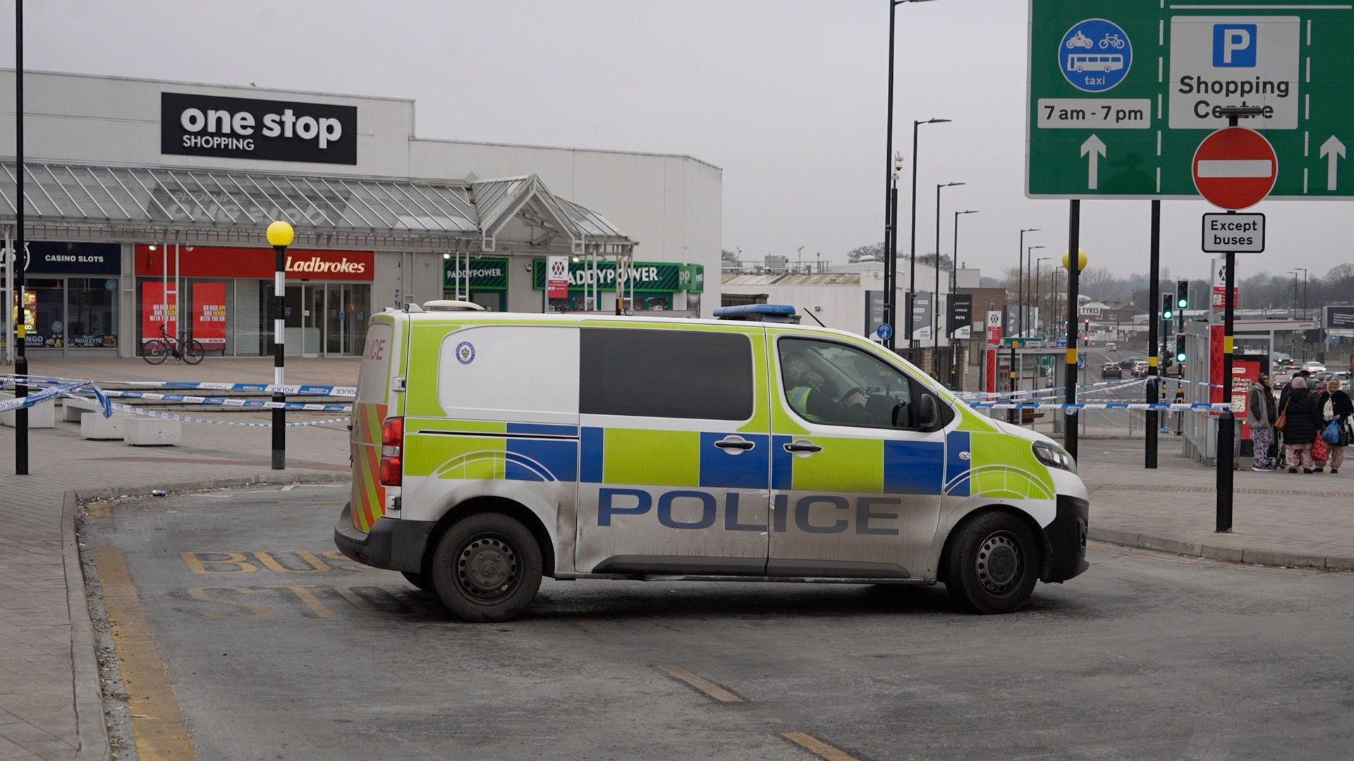 Police van on a street, in front of a cordon around part of Perry Barr. Police tape is wound around lamp-posts, with a shopping area in the background