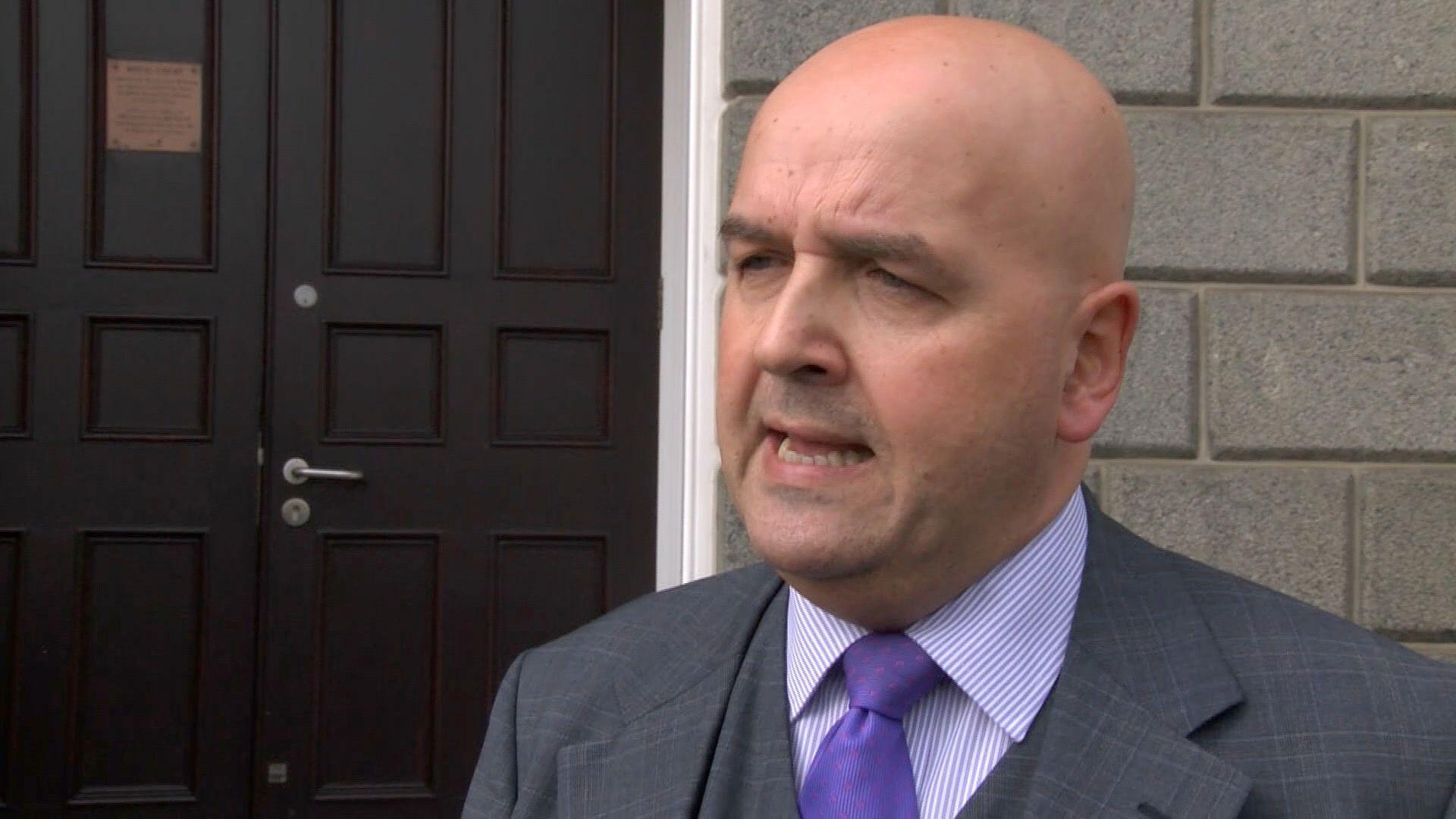 A bald man wearing a suit and tie is seen talking in front of the Guernsey States building - a door and stonework can be seen behind him