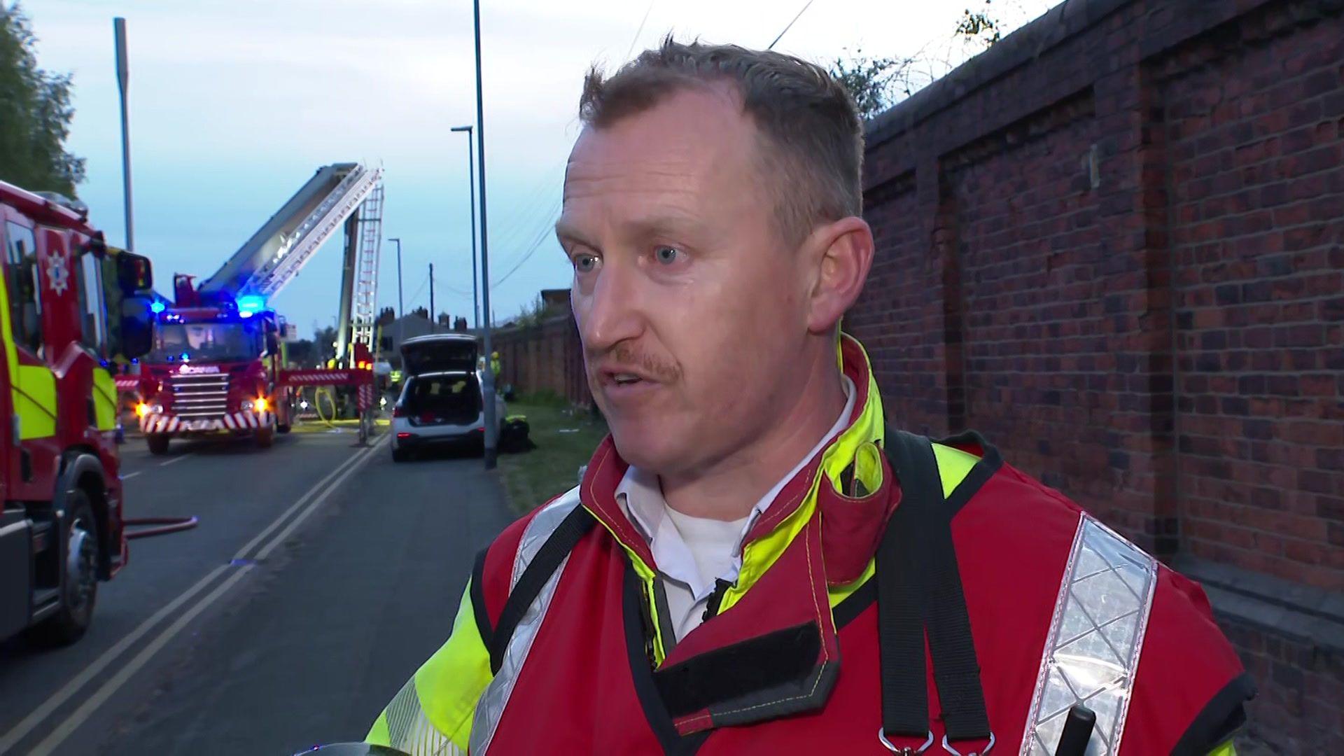 Cheshire Fire And Rescue Service group manager David Rutter speaks to a reporter, off camera. He is standing close to a brick wall. Two fire engines can be seen in the background.