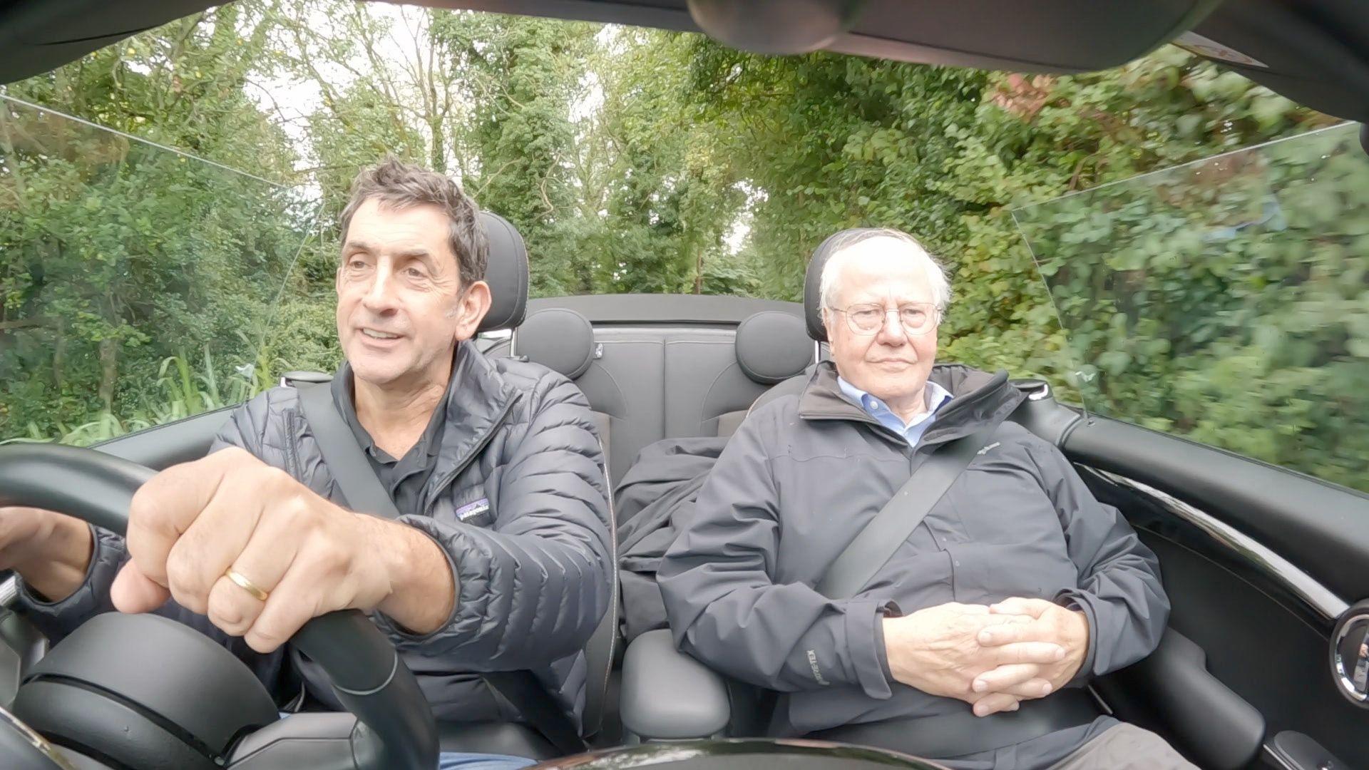 BBC climate editor Justin Rowlatt, dressed in black puffer coat, drives an open-top car down a country lane, beside economist Paul Cheshire in the passenger seat, whose hands are folded across his stomach