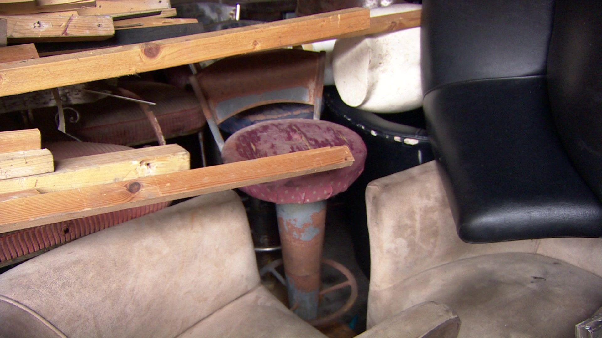 A storage space filled with old, worn-out chairs and wooden planks. The chairs are faded and have rusted metal frames and peeling paint. The wooden planks lie across the chairs.