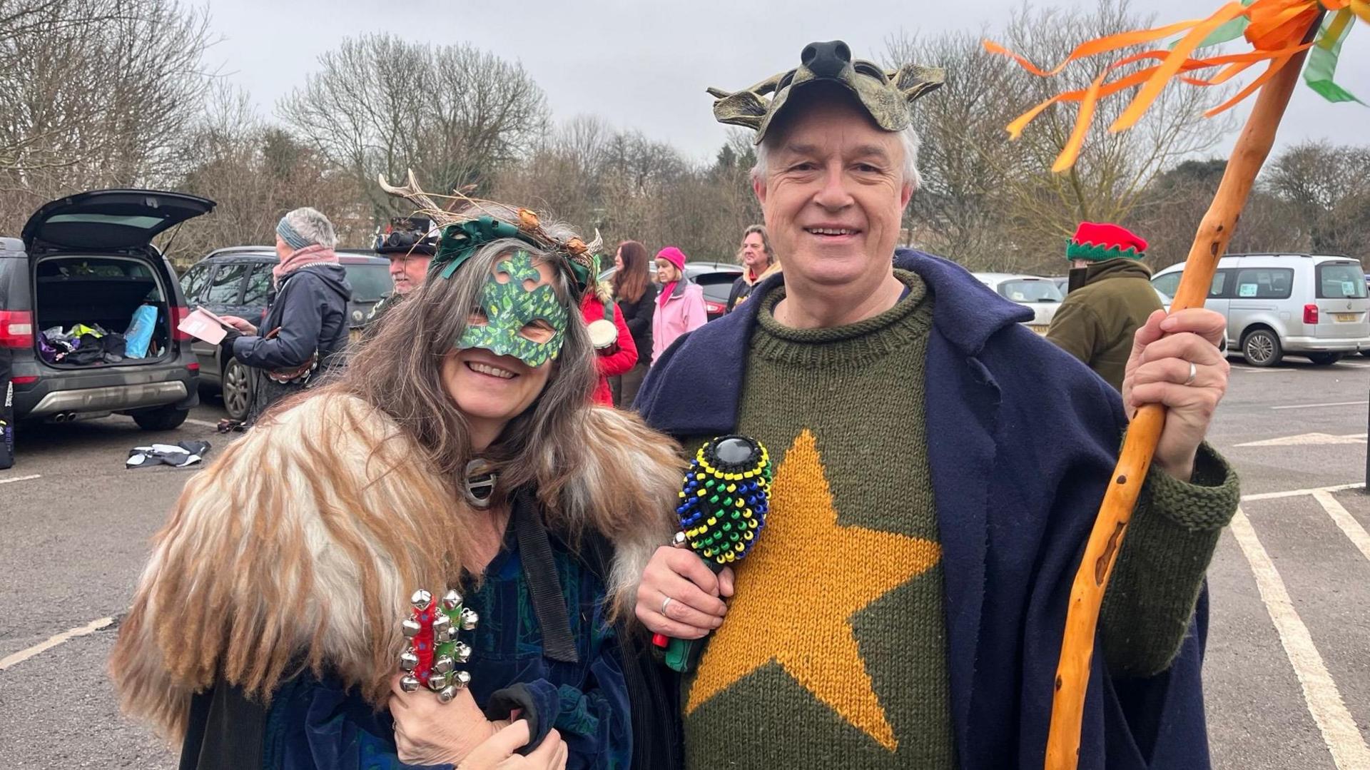 Tracey wears a coat with a large fur collar and a green mask.  Kim wears a deer mask on the top of his on his head and carries a stick with yellow and orange ribbons.  They both hold percussion instruments.