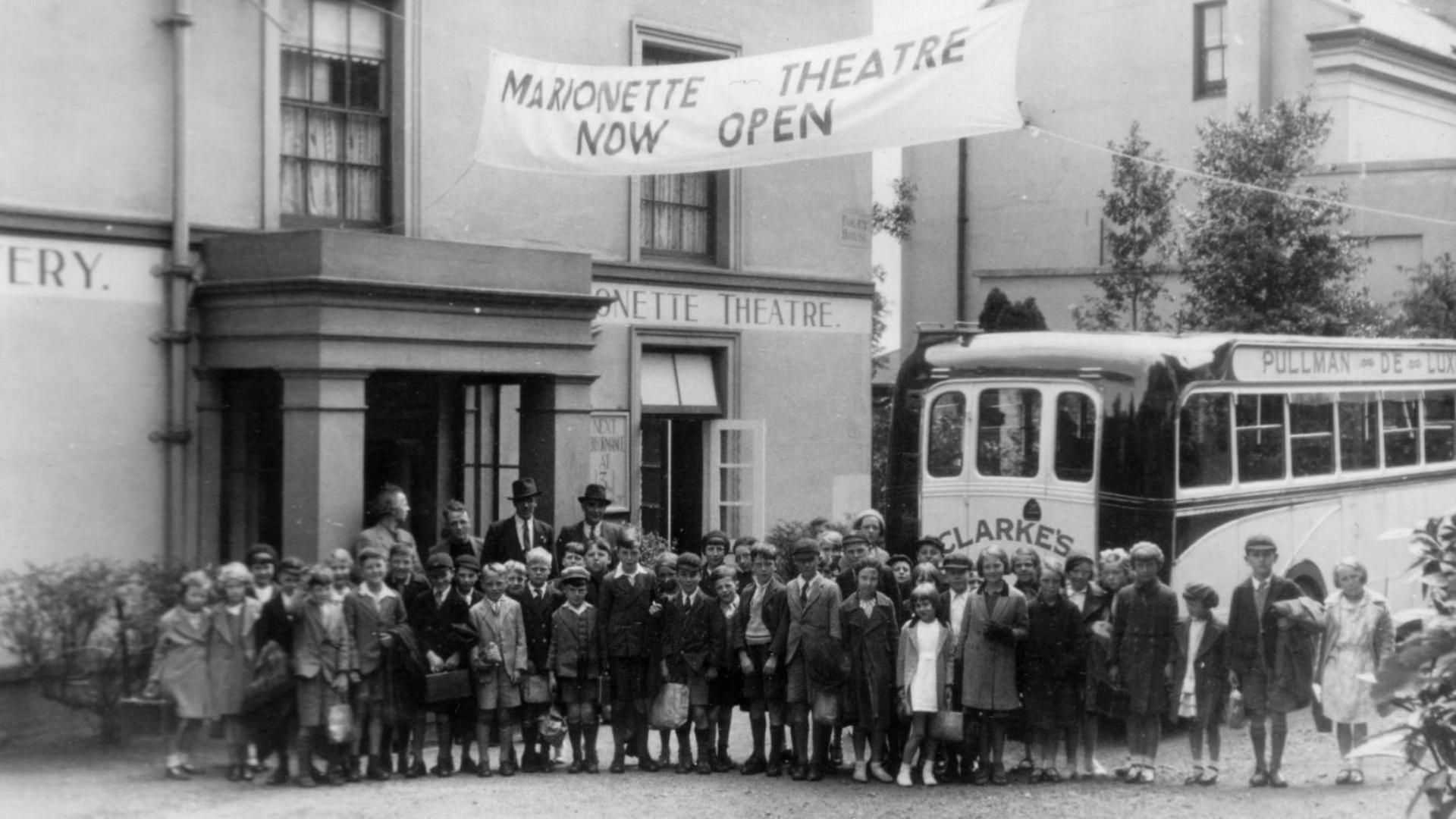 A black and white photo of a large group of schoolchildren in front of a building with a banner attached reading "Marionetter theatre now open". A bus is parked outside the building, behind the children.