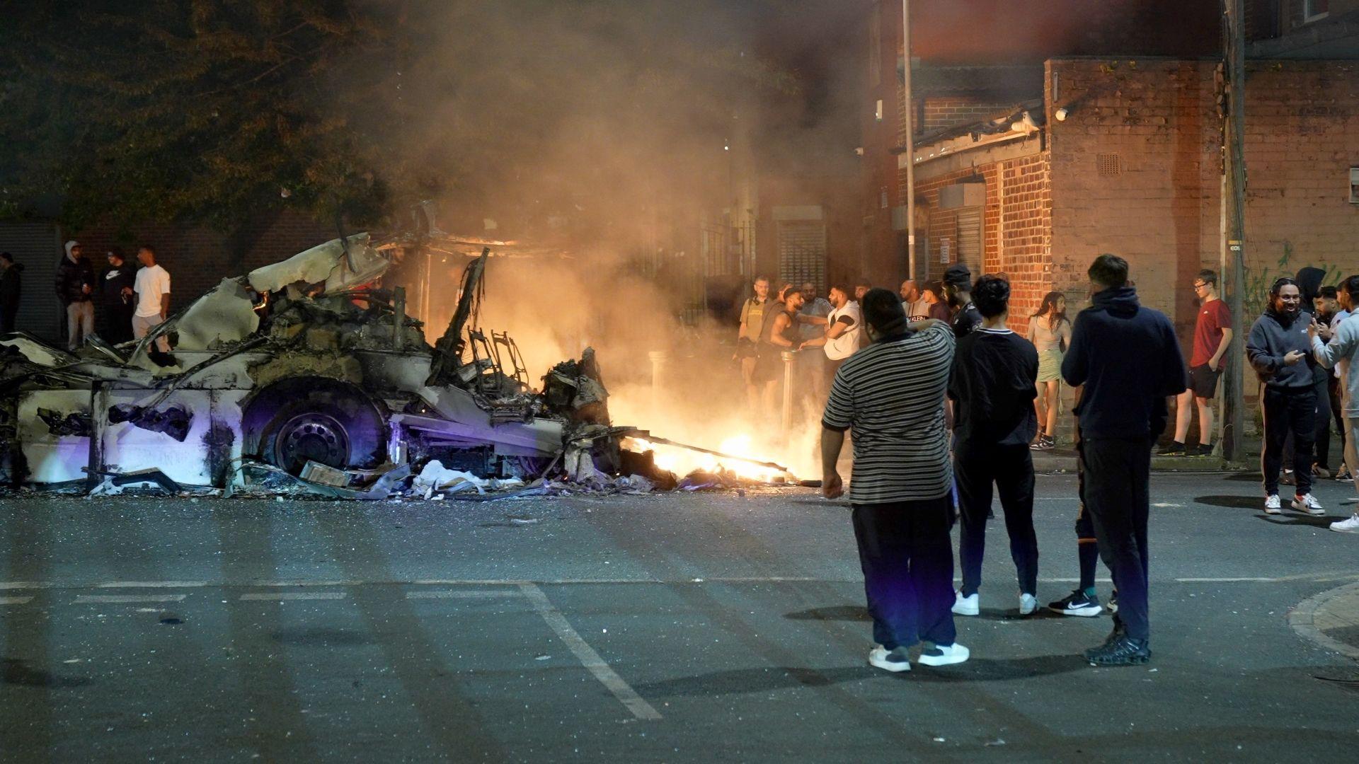 A bus on fire with people standing in front of it