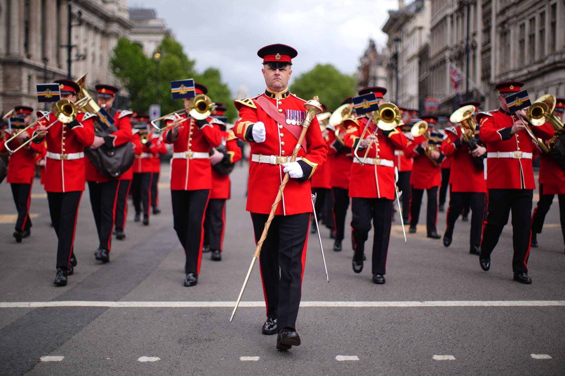 Military members in bright red uniform, carrying gold sceptres and playing brass instruments, walk down the street.