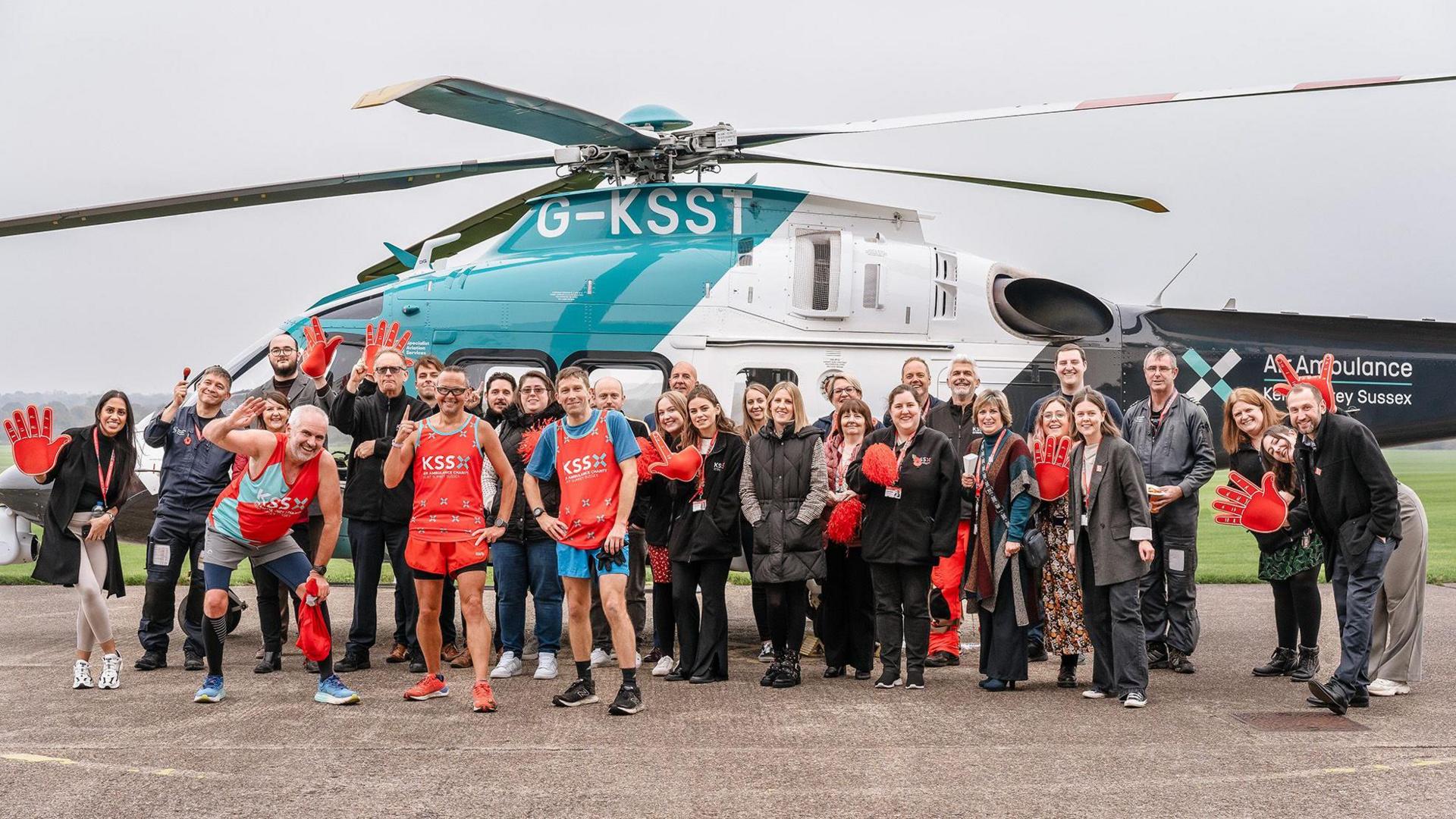 A group photo of Steve Wolfe and the KSS team. There are around 30 people in the photo all smiling at the camera. Behind the group is a KSS helicopter. 