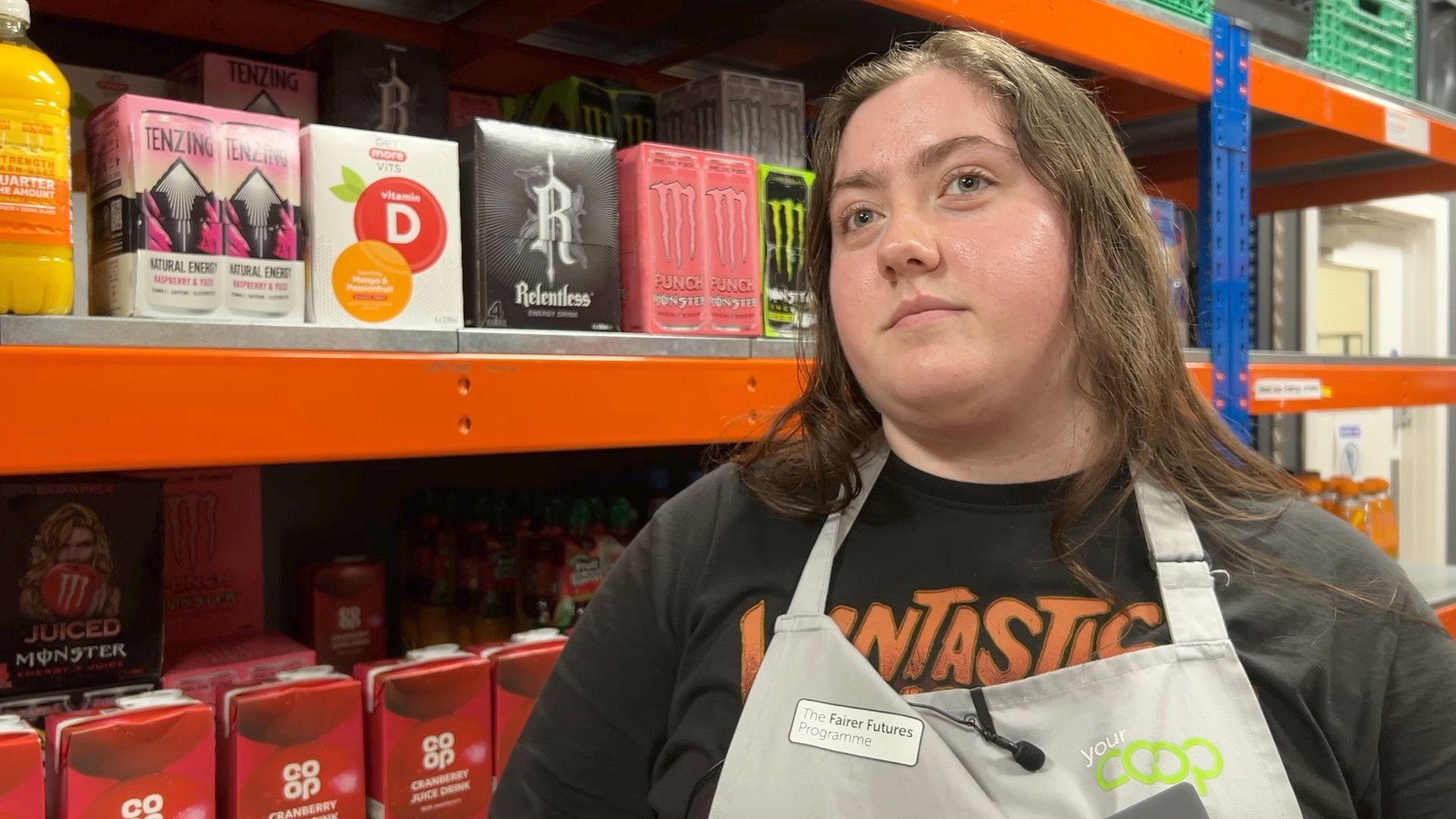 A woman stands in front of drinks products on a supermarket shelf. She has straight brown hair and is wearing a black top and a grey apron. The apron has a green "Your Co-op" logo on it. 