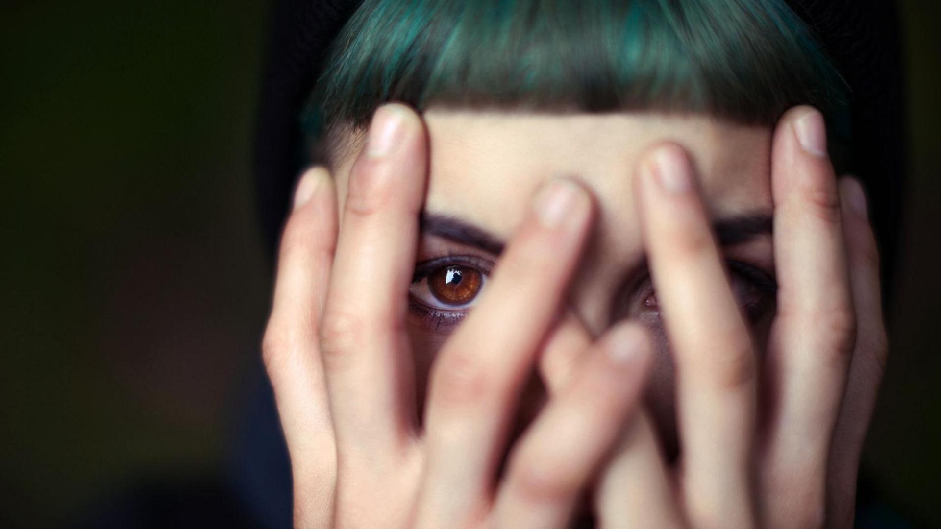 A woman with brown eyes and dyed green hair peeks through her fingers