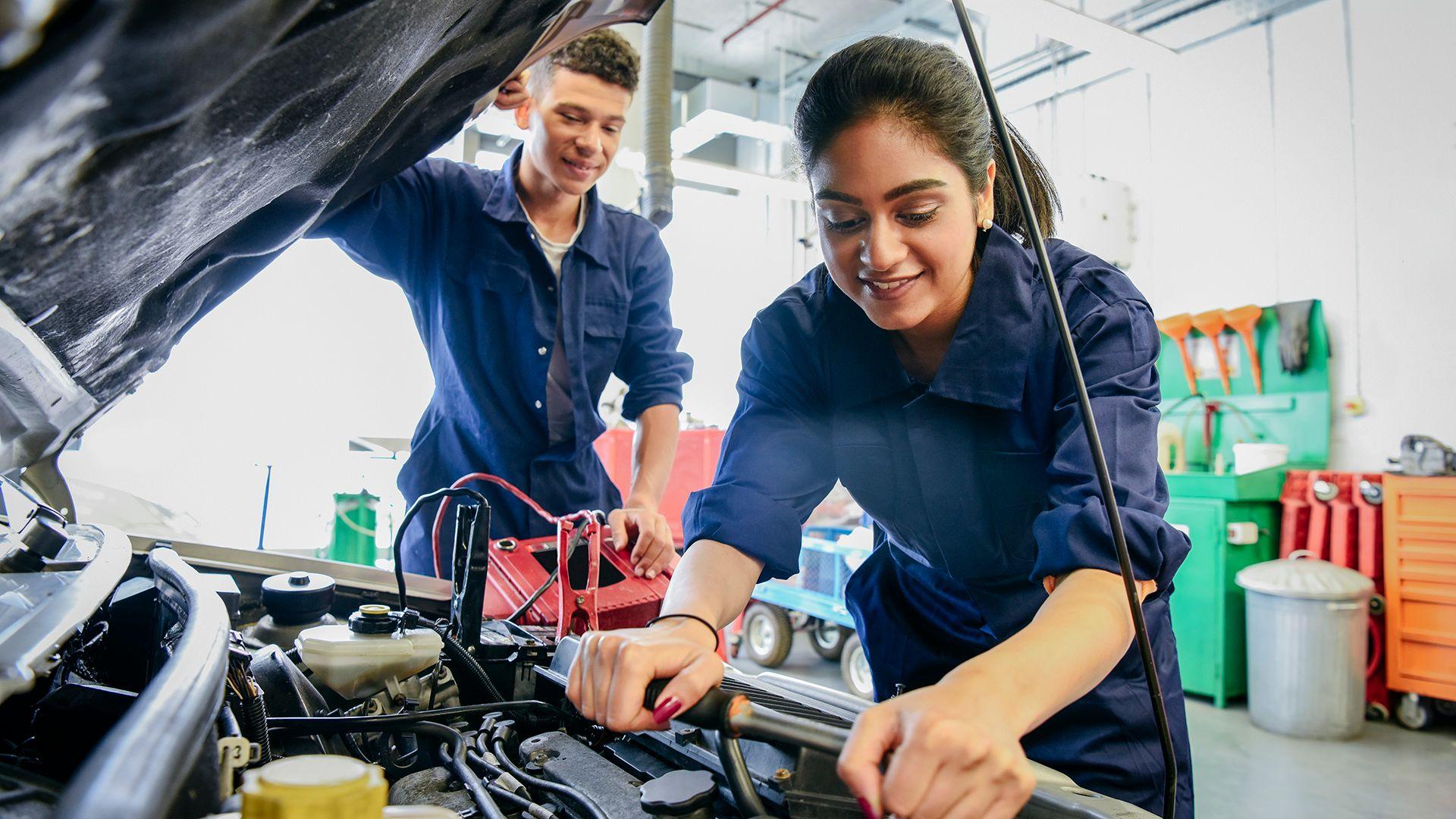 Stock photo shows a young person wearing blue overalls adjusts an engine in the bonnet of a car with it's panel up, while another worker looks on in the background, in a mechanic shop.