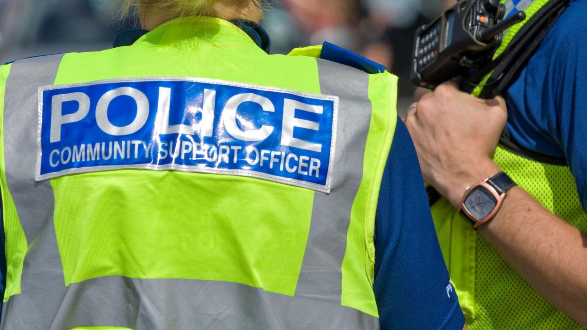 A woman wearing a fluorescent yellow jacket with white writing on the back set against a blue backdrop which reads Police Community Support Officer. She is stood next to a make who is also wearing a fluorescent yellow jacket with a radio device attached to the jacket on his chest. 