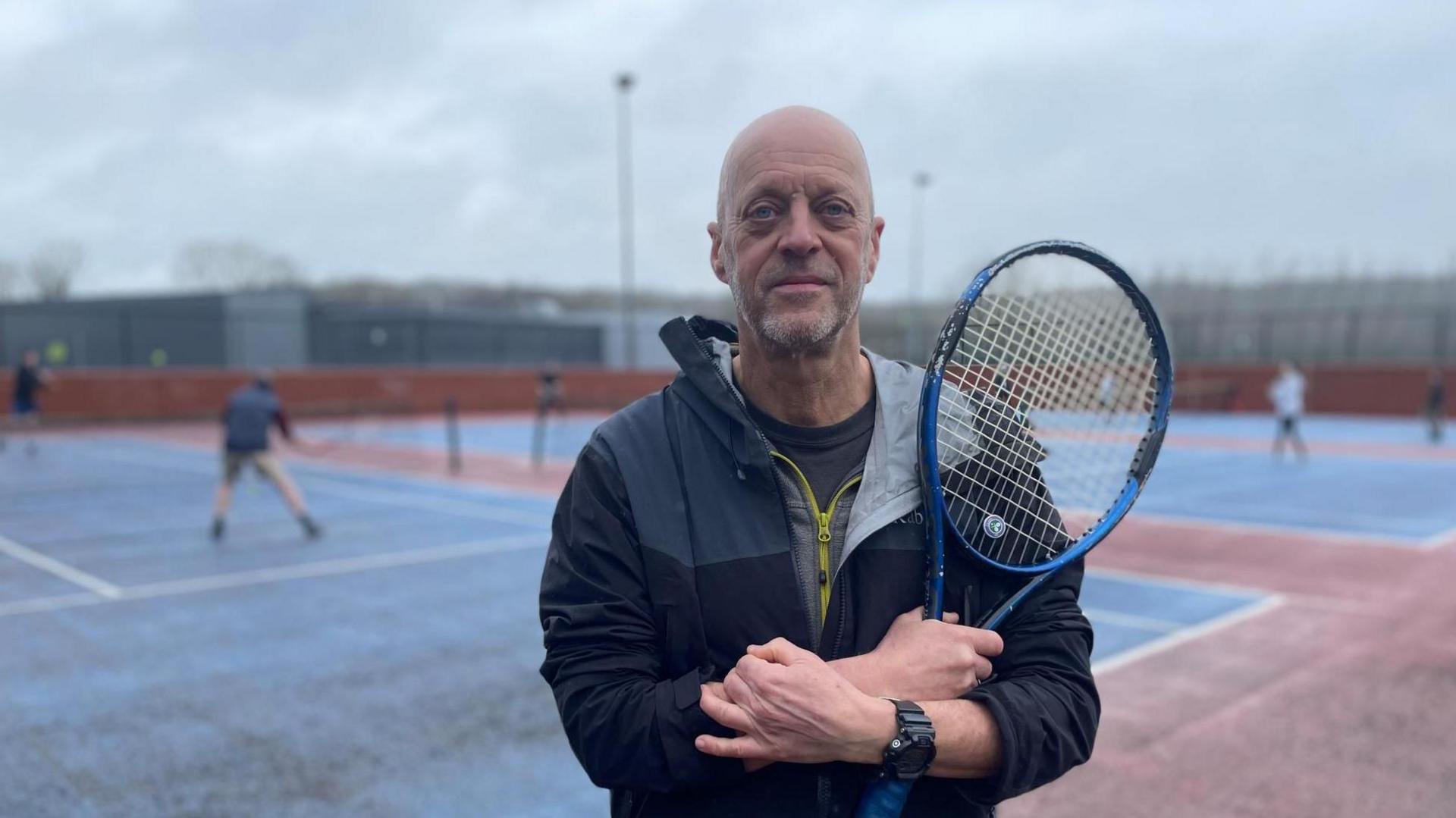 A man stands with folded arms holding a tennis racquet next to an old dirty tennis court