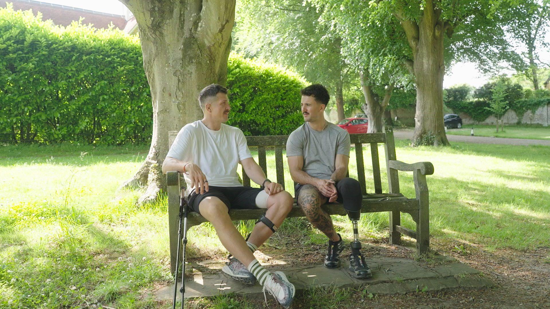 Two individuals, one with a prosthetic leg, sitting on a wooden bench in a park, surrounded by trees.