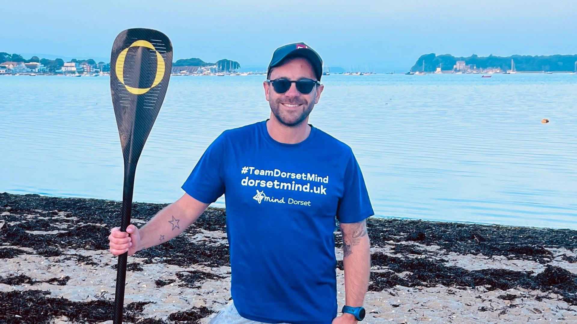 David Haze standing on a beach with Brownsea Island and the entrance to Poole Harbour in the background. He is smiling at the camera and holding a paddle in his right hand. He's wearing sunglasses, a cap and a blue t-shirt bearing the Dorset Mind logo