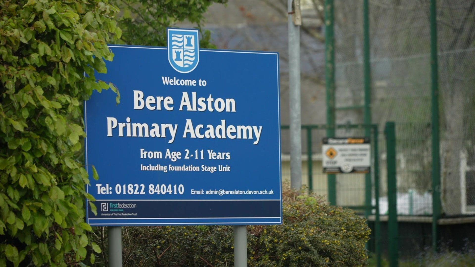 A blue sign with the words 'Welcome to Bere Alston Primary Academy'. There is bush to the left and a green fence in the background on the right.