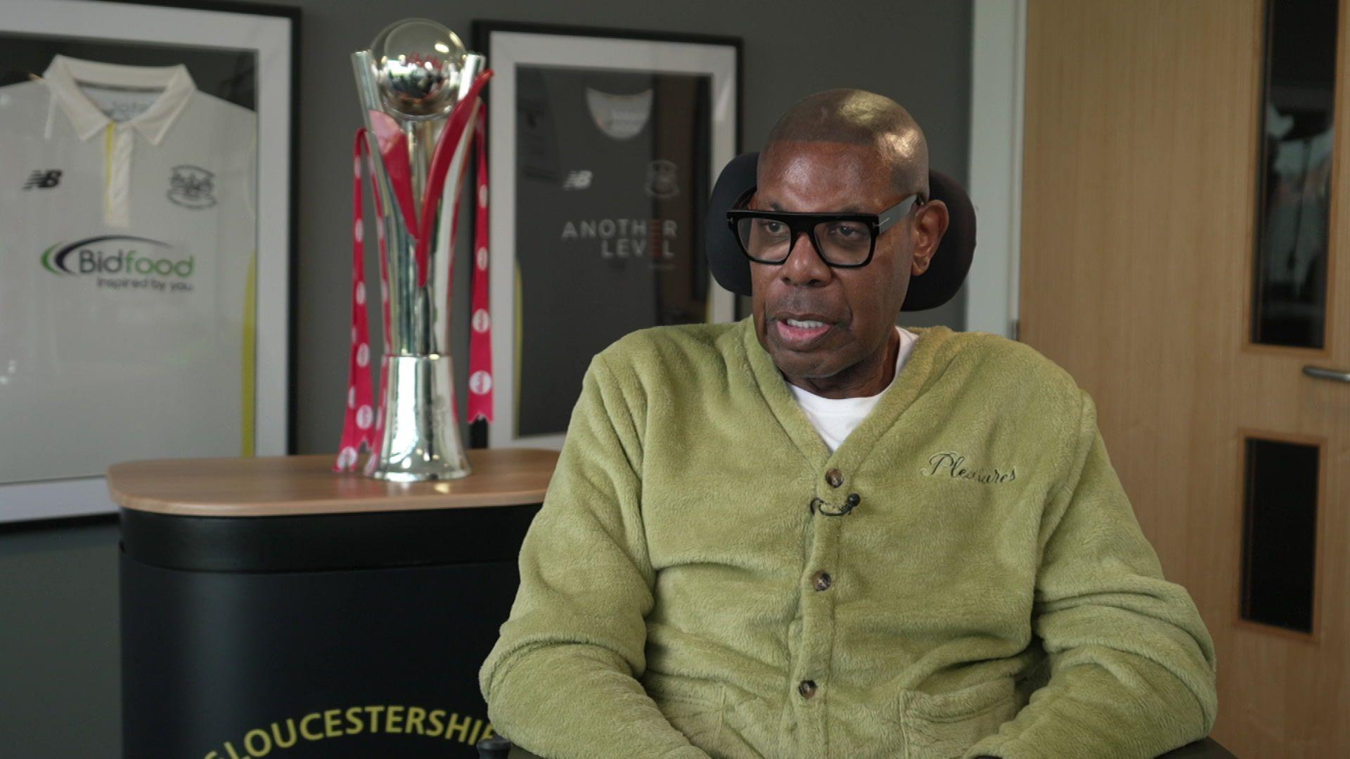 David 'Syd' Lawrence. He is wearing black spectacles and a green cardigan with a white t-shirt underneath. A trophy and framed cricket shirts are behind him