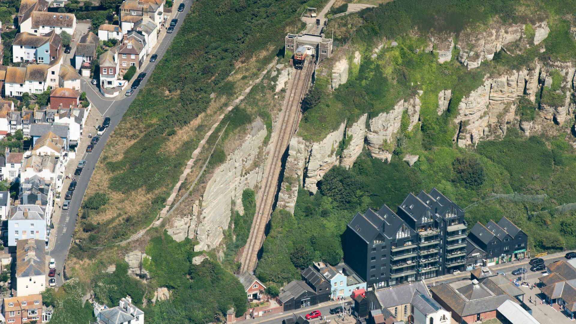 An aerial view of the East Hill Cliff Railway in Hastings. There are houses on the left and blocks of flats on the right at the base of the cliff. Most of the cliff is covered in green plants. Some of the grey cliff face is visible