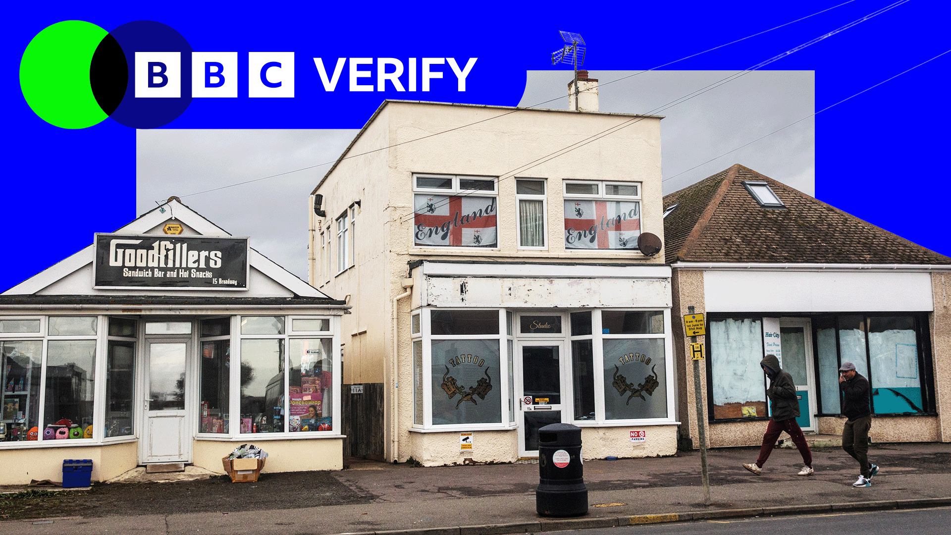 A photo of three small commercial buildings in Jaywick, near Clacton-on-Sea in Essex. The left unit is a sandwich shop named “Goodfillers,” the central building displays England flags in the windows above a tattoo studio, and the shop on the right has papered-over windows and a faded sign. Two men in are walking past, wearing dark clothing. The sky is grey and overcast. A BBC Verify logo is superimposed on the upper portion of the image with a blue border.