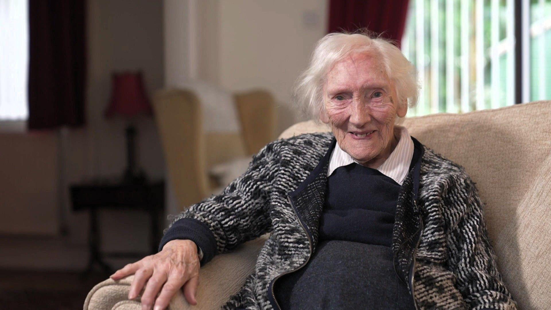 Menyw mewn oed gyda gwallt llwyd a gwyn, yn gwenu tuag at y camera. Mae hi'n eistedd ar sofa lliw golau mewn lolfa taclus.