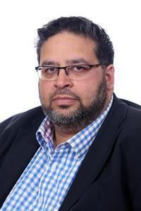 A dark-haired, bearded man wearing glasses, a blue-checked shirt and black jacket