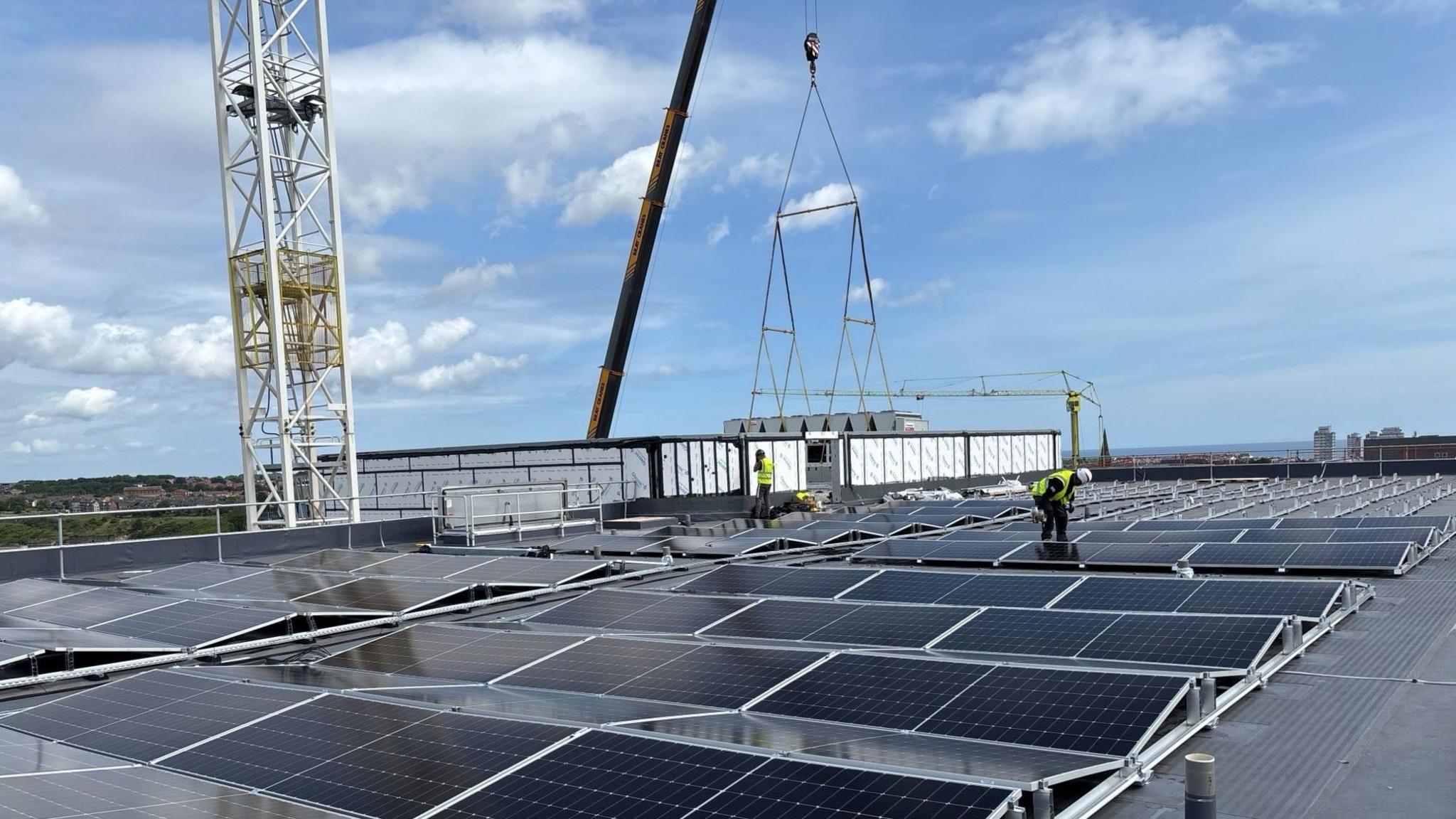 Solar panels on top of the new hospital building. Two men in high-viz vests and hard hats are working on the roof, with a crane in the background.
