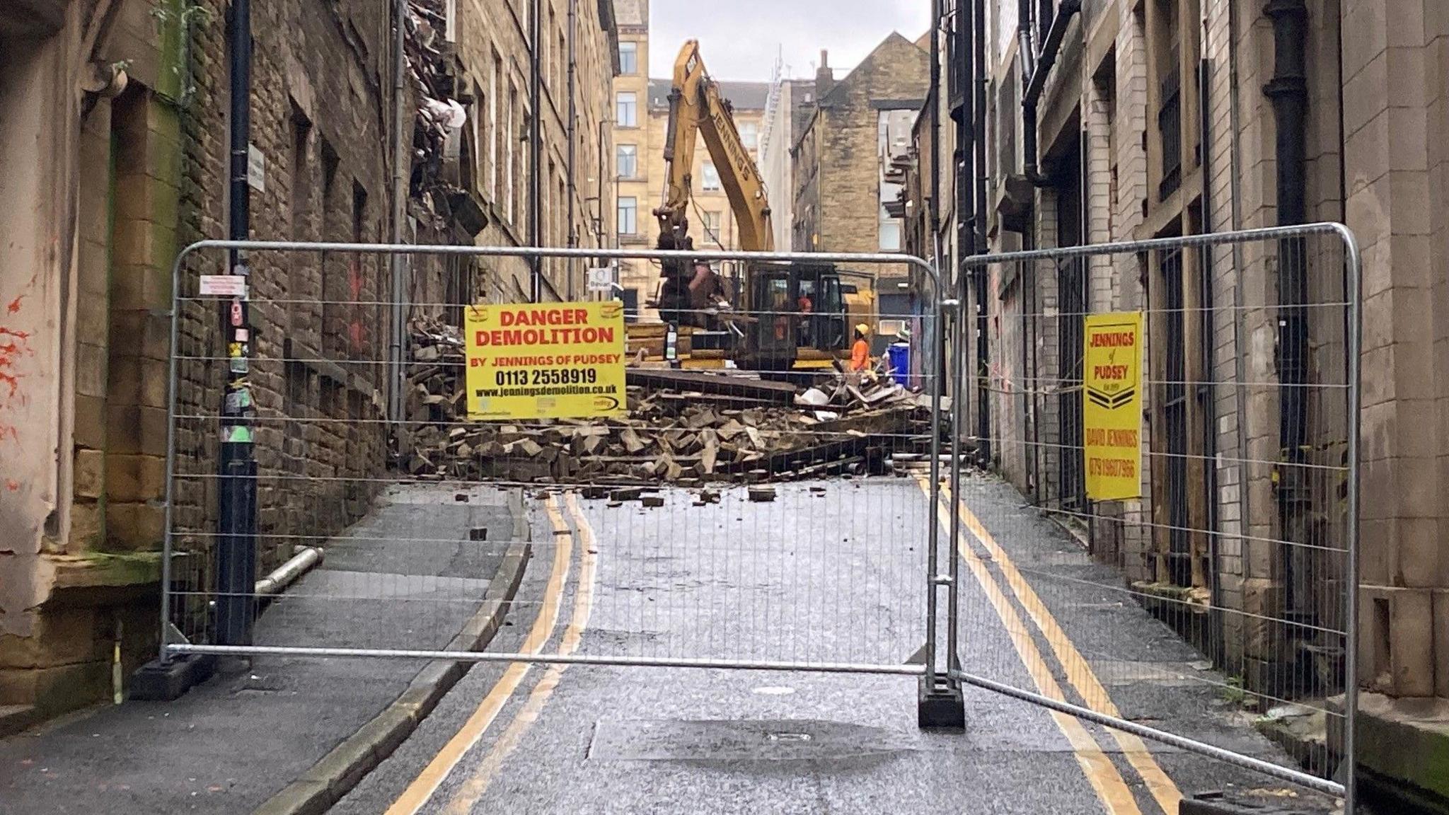 A yellow mechanical excavator working behind metal fencing on street with double yellow parking lines. 
