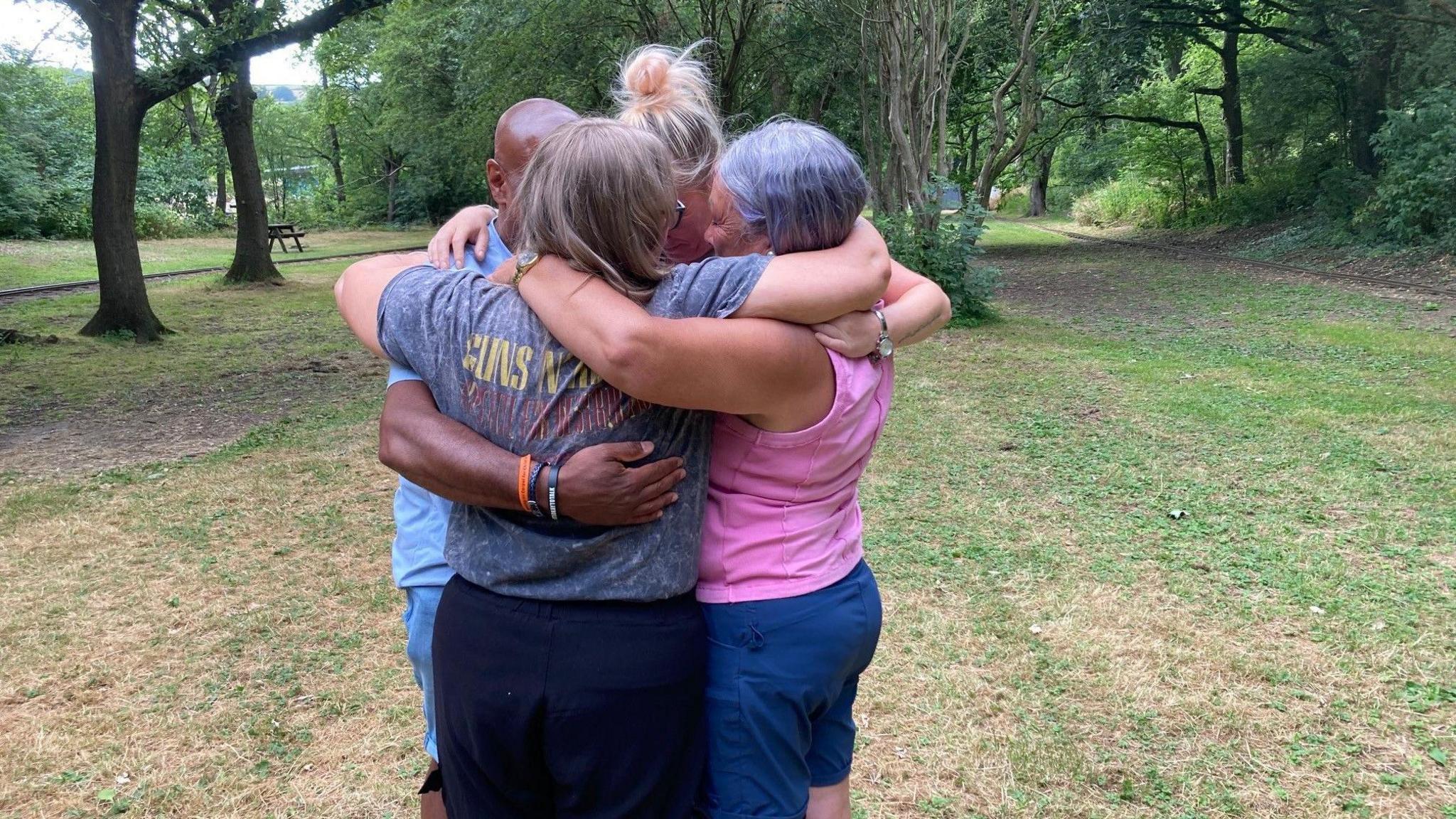 Four adults hug each other in a park. Three are women and there is one man.
