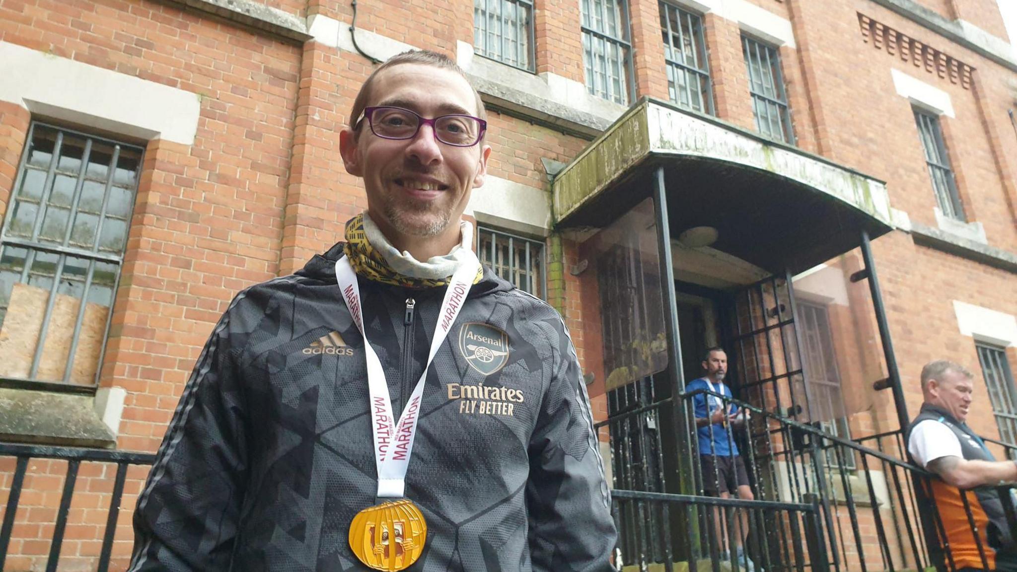 David Andrewartha. He has short brown hair, has purple glasses on, a black tracksuit top and medal shaped like a pumpkin around his neck. Behind him is a red brick building with metal grates on the window